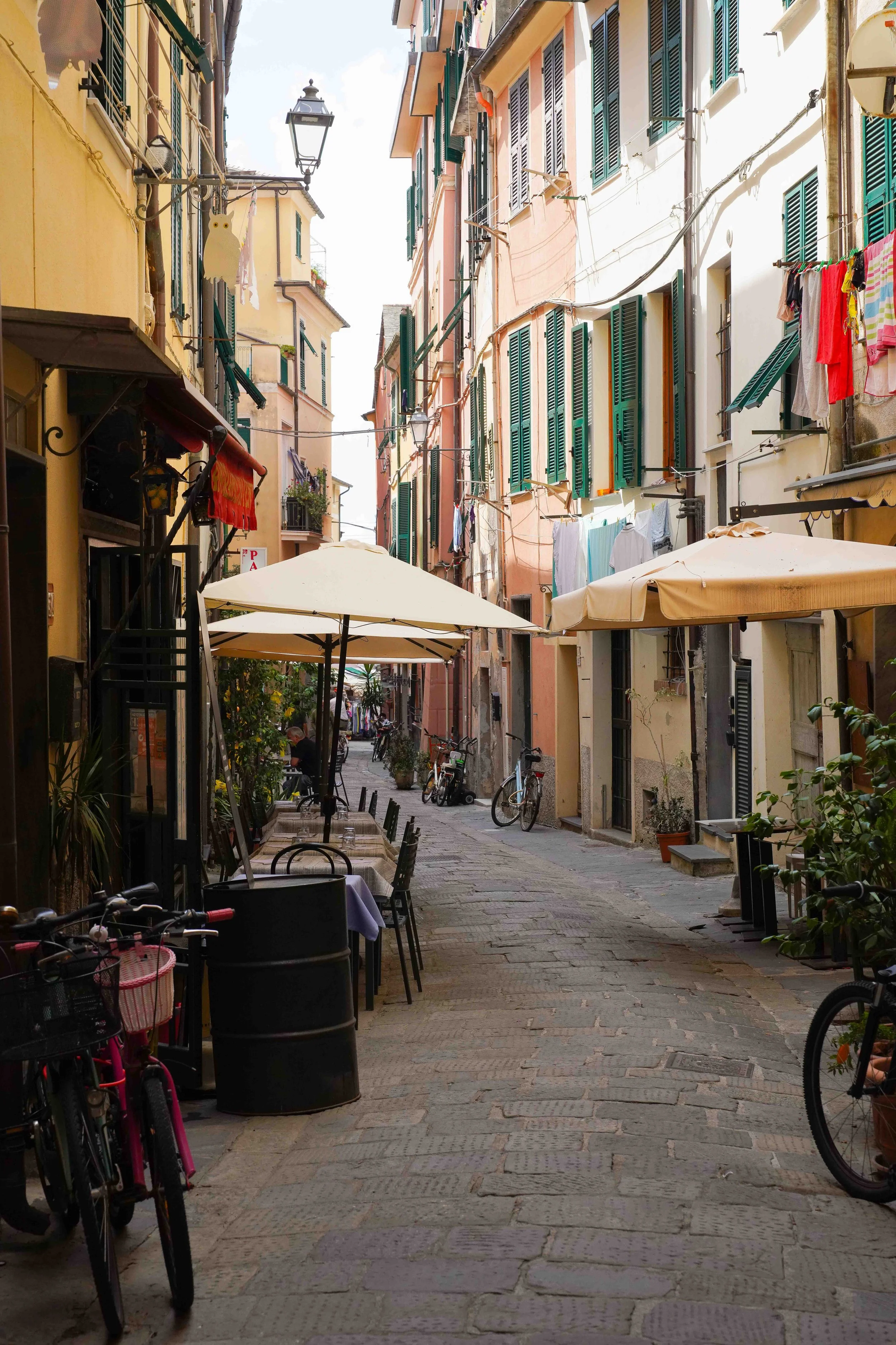 A narrow, cobblestone street in Italy with outdoor café tables, bicycles parked along the sides, colorful buildings with green shutters, hanging laundry, and umbrellas providing shade.