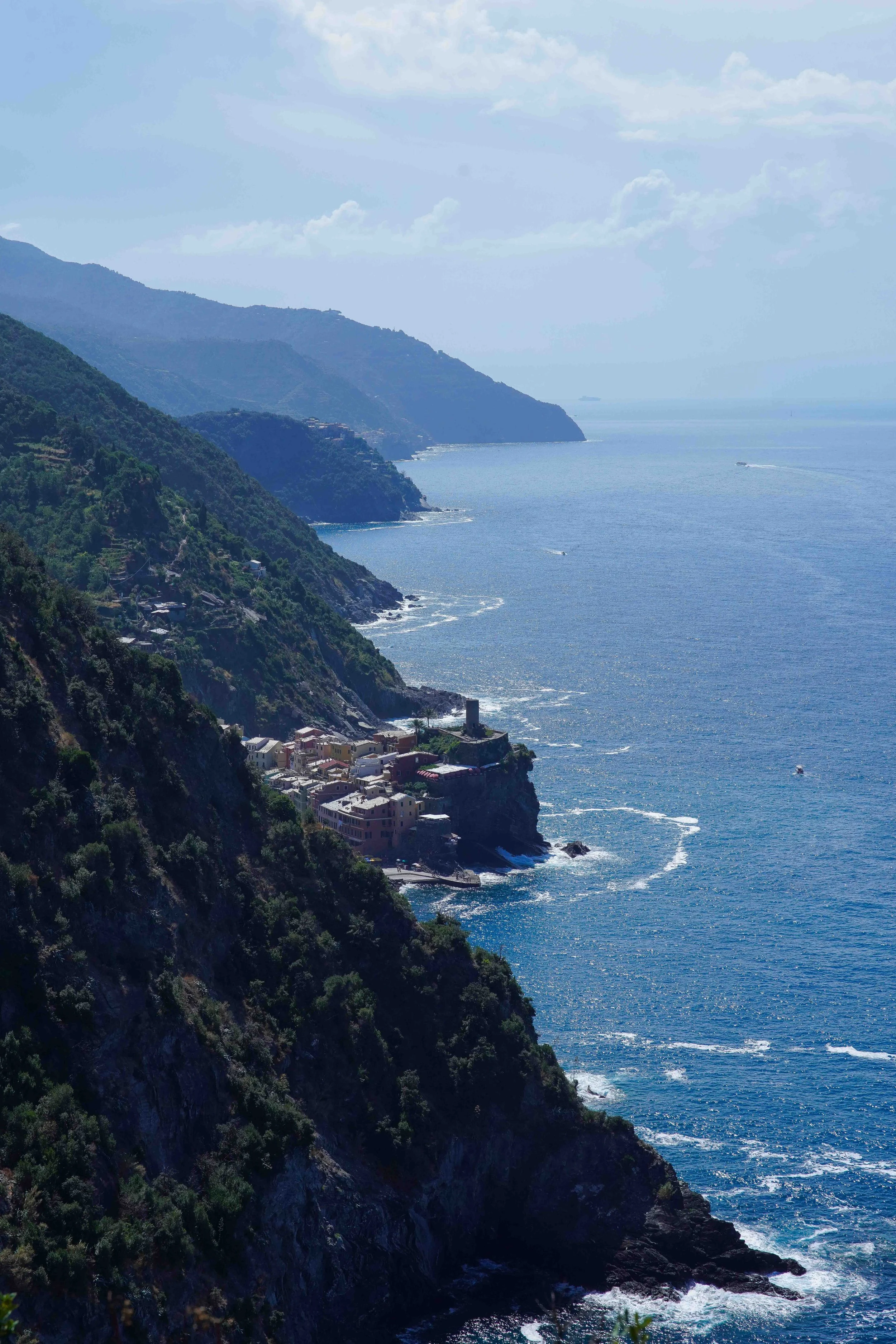 Blue Trail in Cinque Terre offering view of cliffs with lush green vegetation and Vernazza at the base, overlooking the ocean with boats and a ship in the distance under a blue sky with scattered clouds.