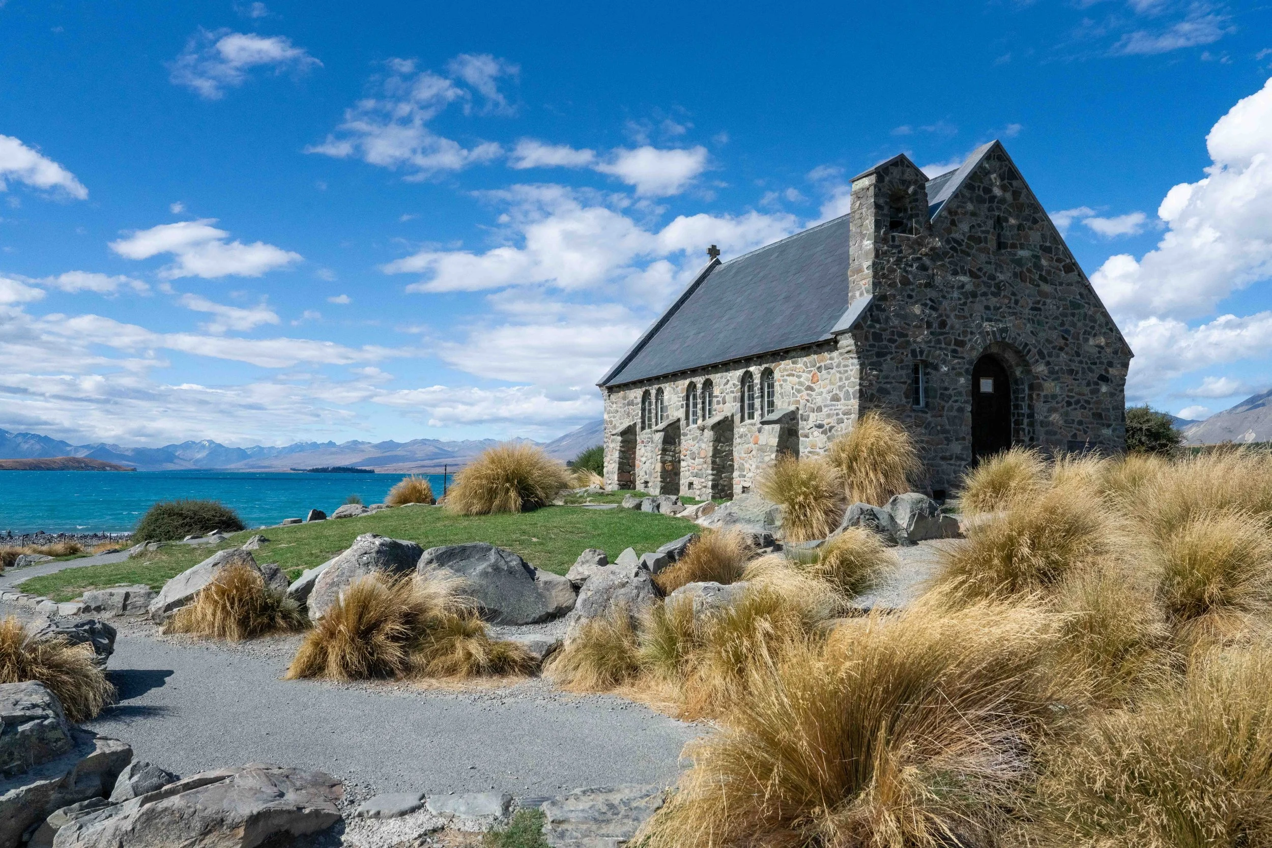 Stone church in Lake Tekapo, New Zealand, with mountains in the background under a partly cloudy sky.