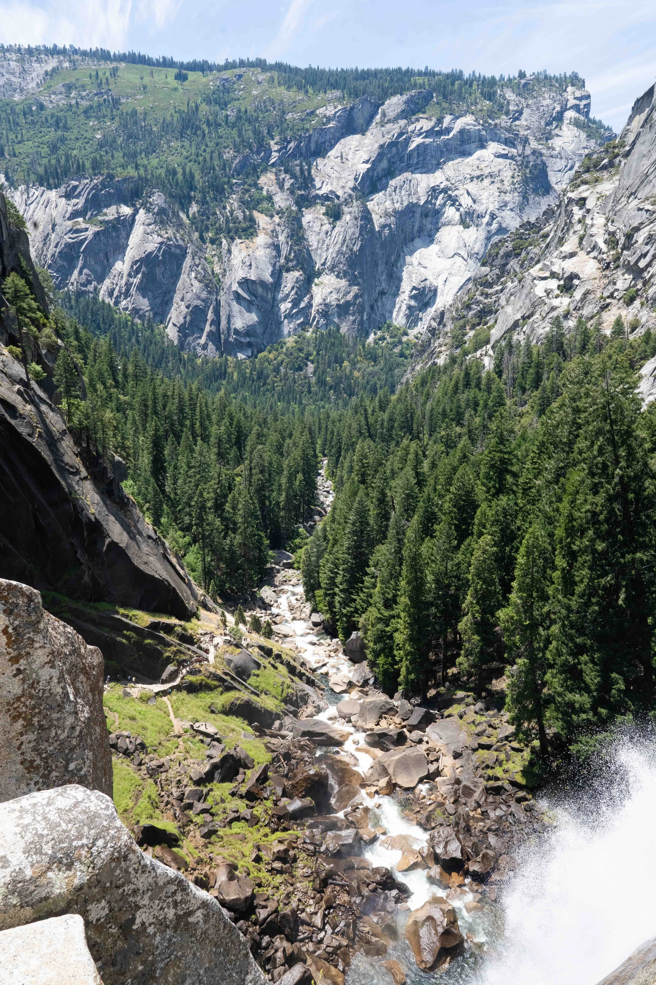 Scenic view in Yosemite of Vernal Falls' mountain landscape with large granite cliffs, lush green forest in the foreground, and a stream flowing through the valley.
