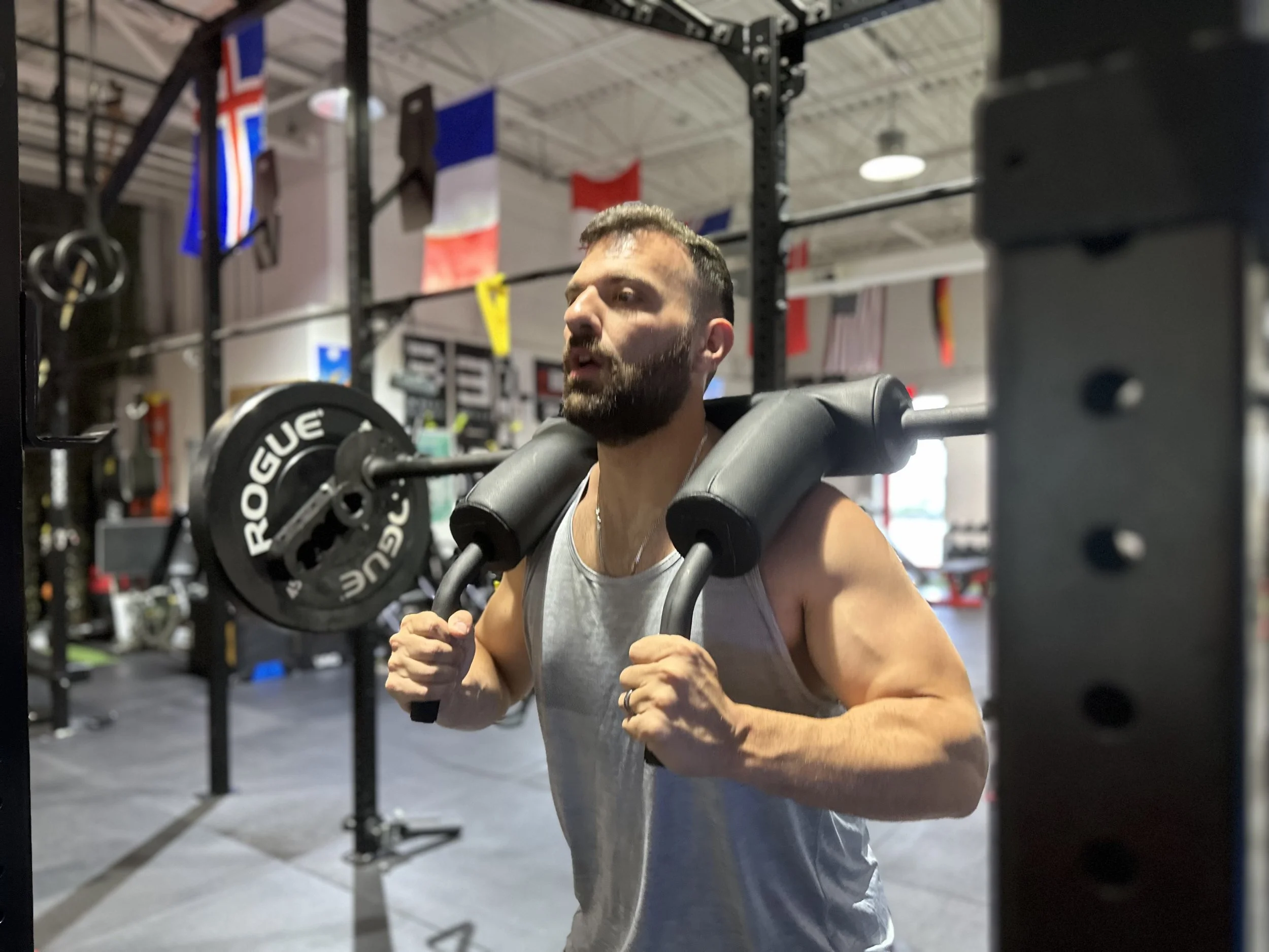 A man with a beard and short hair lifting weights in a gym, with various flags hanging from the ceiling in the background.