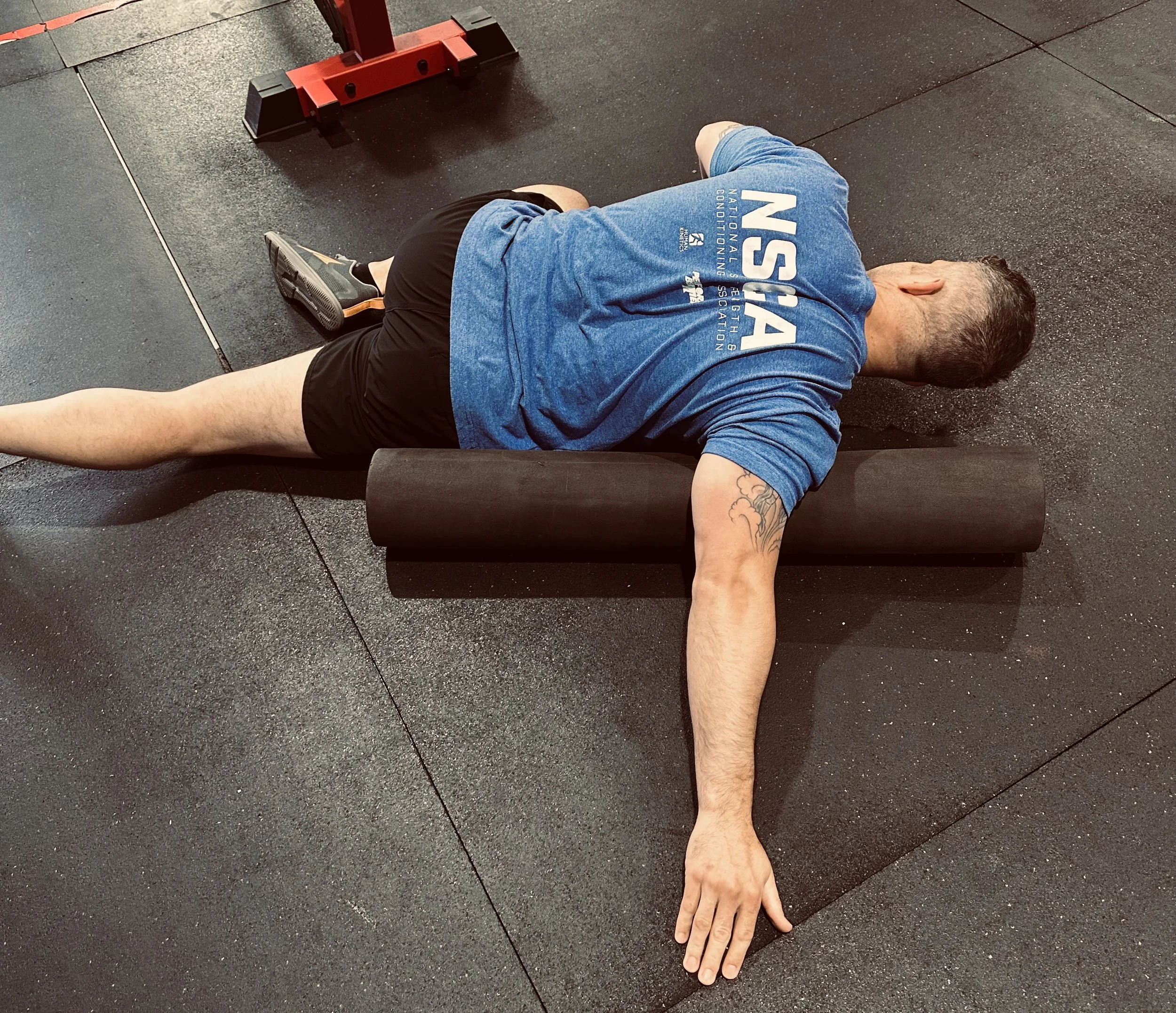A person lying on the gym floor with their head turned to the side, wearing a blue T-shirt and black shorts, next to black gym mats and a red weightlifting stand.