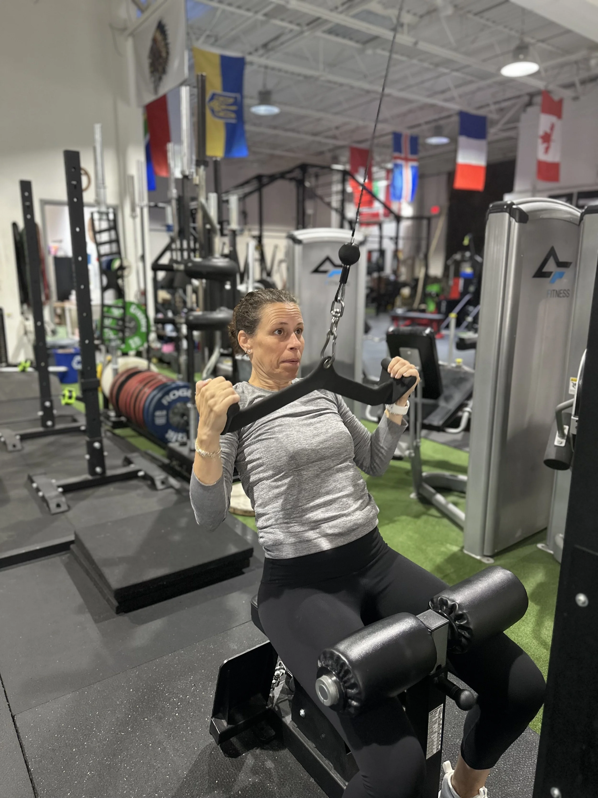 A woman using a seated pulldown machine at a gym, with various flags hanging from the ceiling in the background.