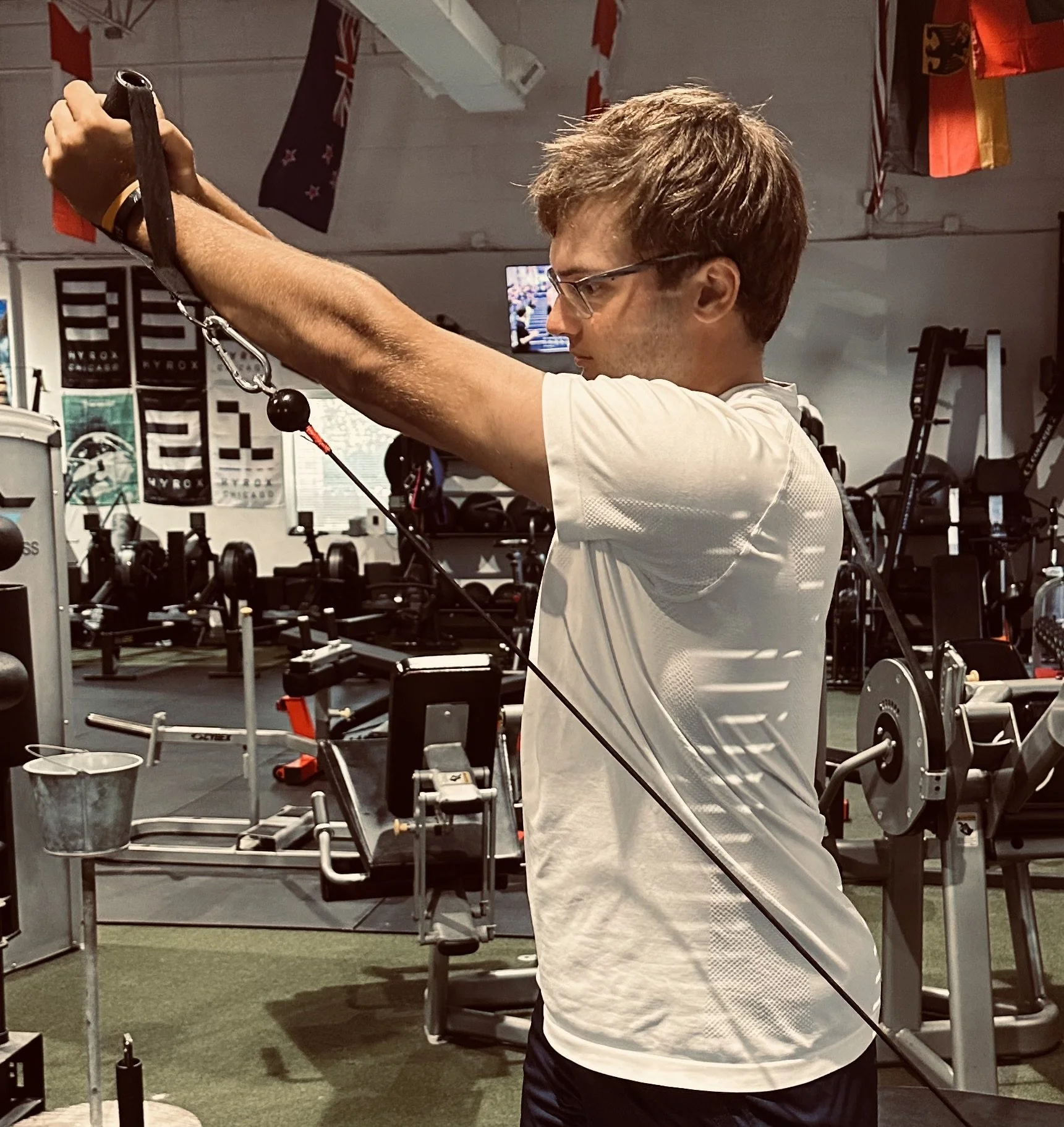 A man wearing glasses and a white athletic shirt is working out in a gym. He is holding a cable attachment with both hands, extending his arms forward. The gym has various equipment, decorations, and flags hanging from the ceiling.