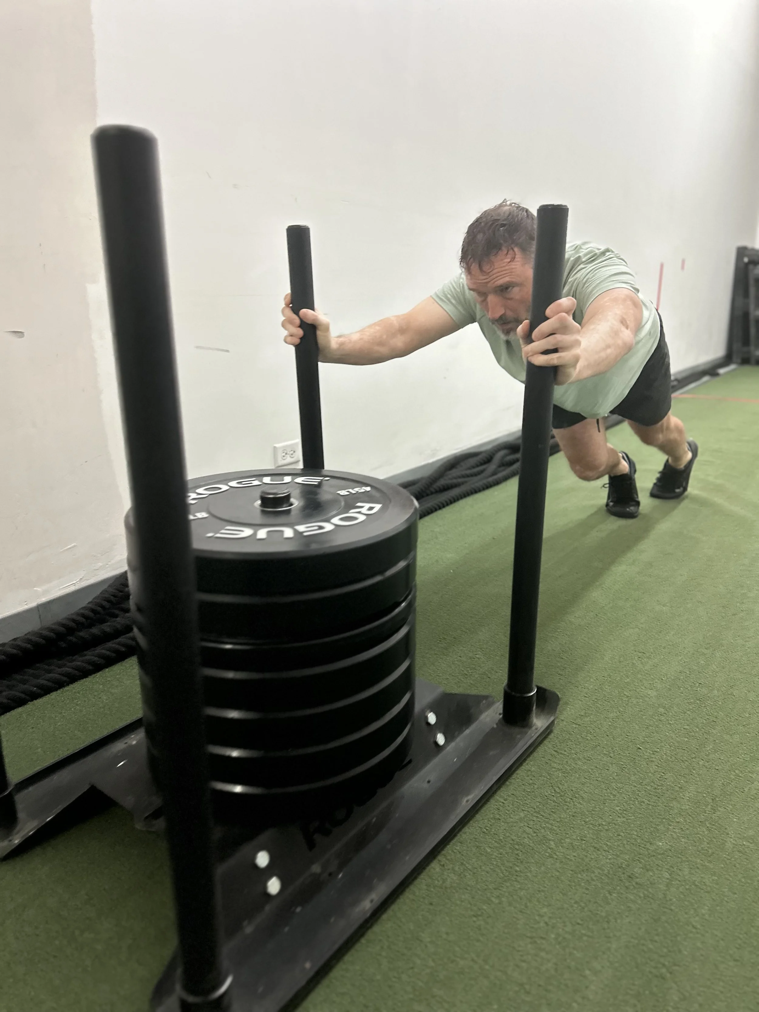 A man in workout clothes pushing a sled loaded with weight plates in a gym.