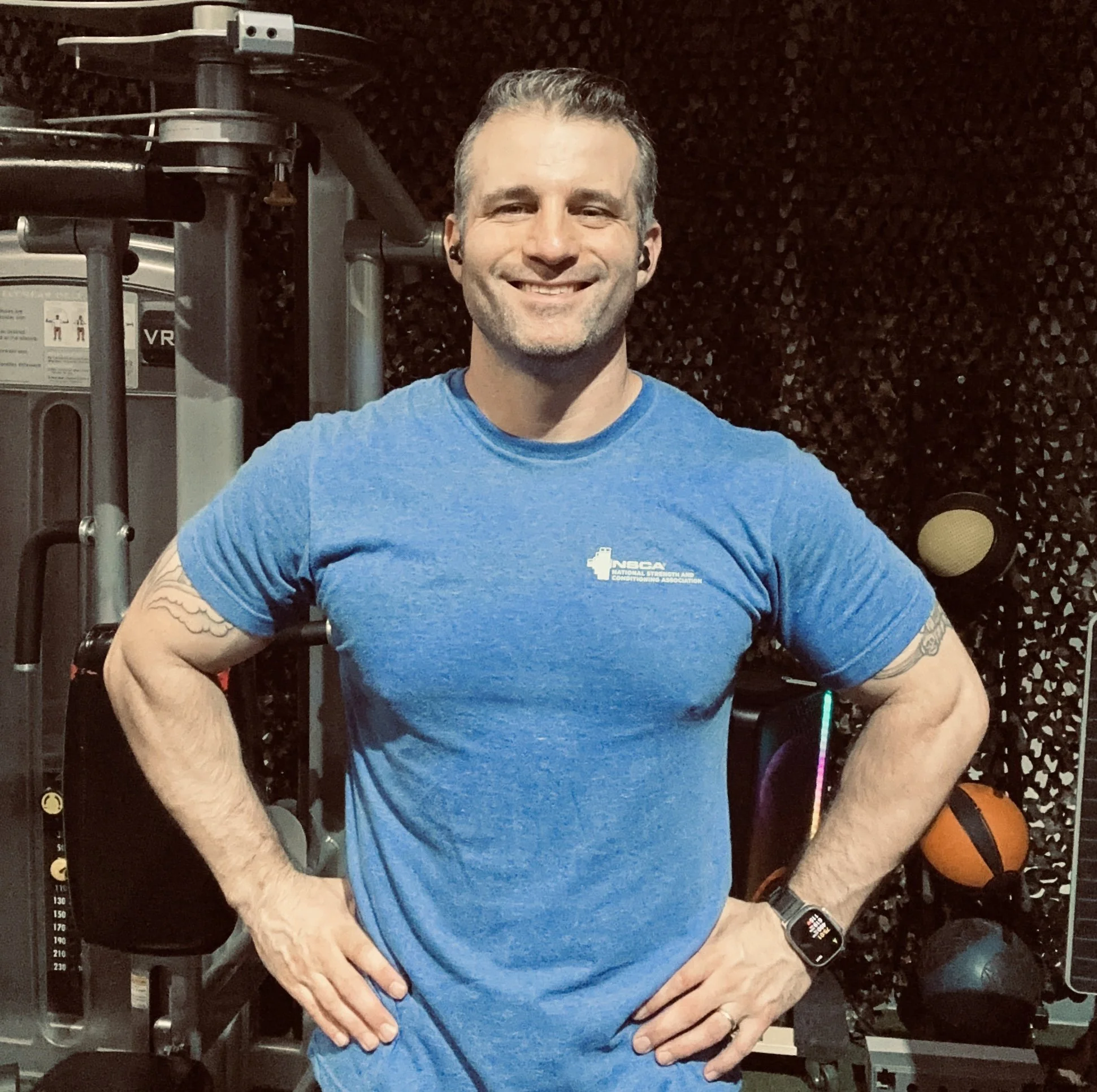 A man with short gray hair, smiling, wearing a blue t-shirt and a smartwatch, standing in a gym with workout equipment and weights in the background.