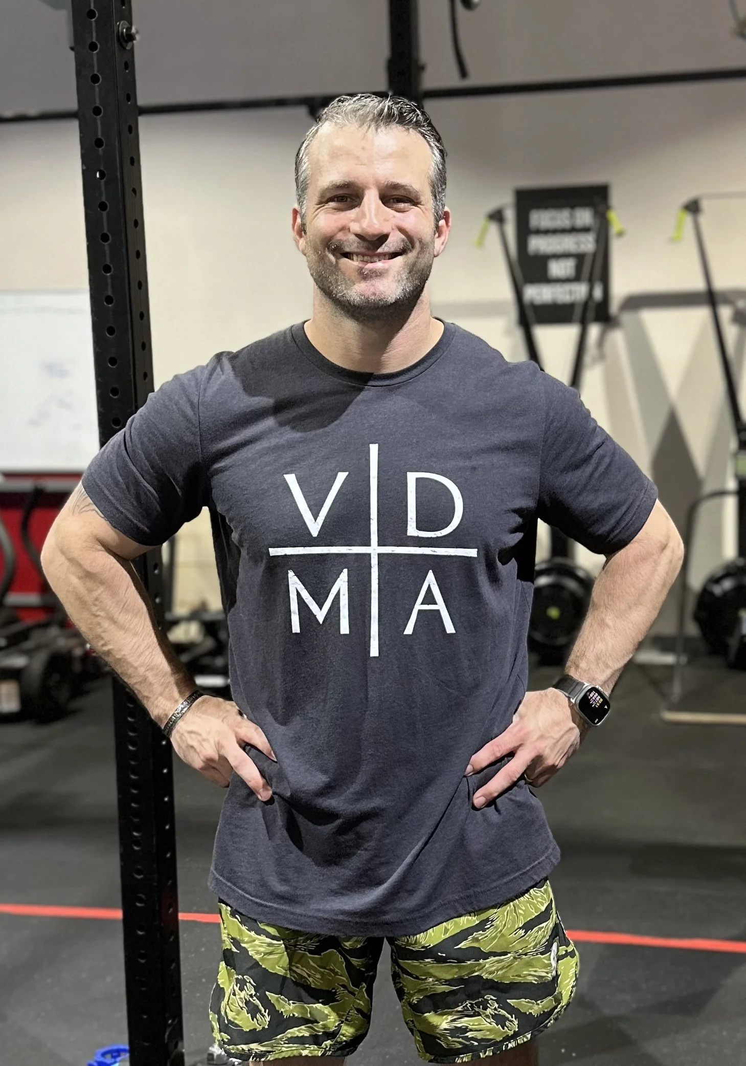 Man standing in a gym, smiling, wearing a gray t-shirt with "VD M A" printed on it, yellow camouflage shorts, and a wristwatch, with gym equipment in the background.