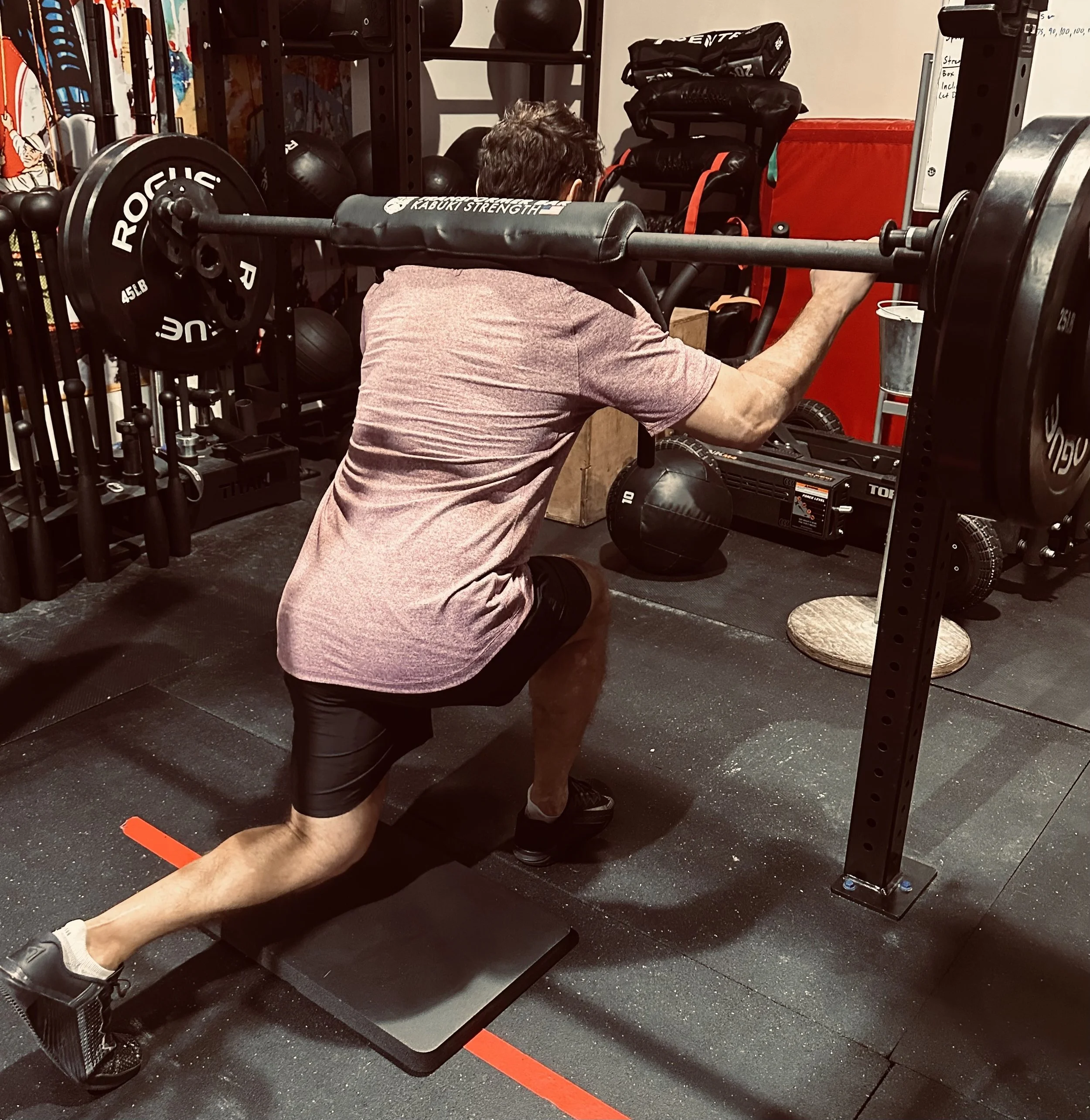 A person with dark hair and wearing a pinkish t-shirt and black shorts is doing a deadlift exercise in a gym. The person is lifting a barbell loaded with weight plates while standing on a black rubber floor.
