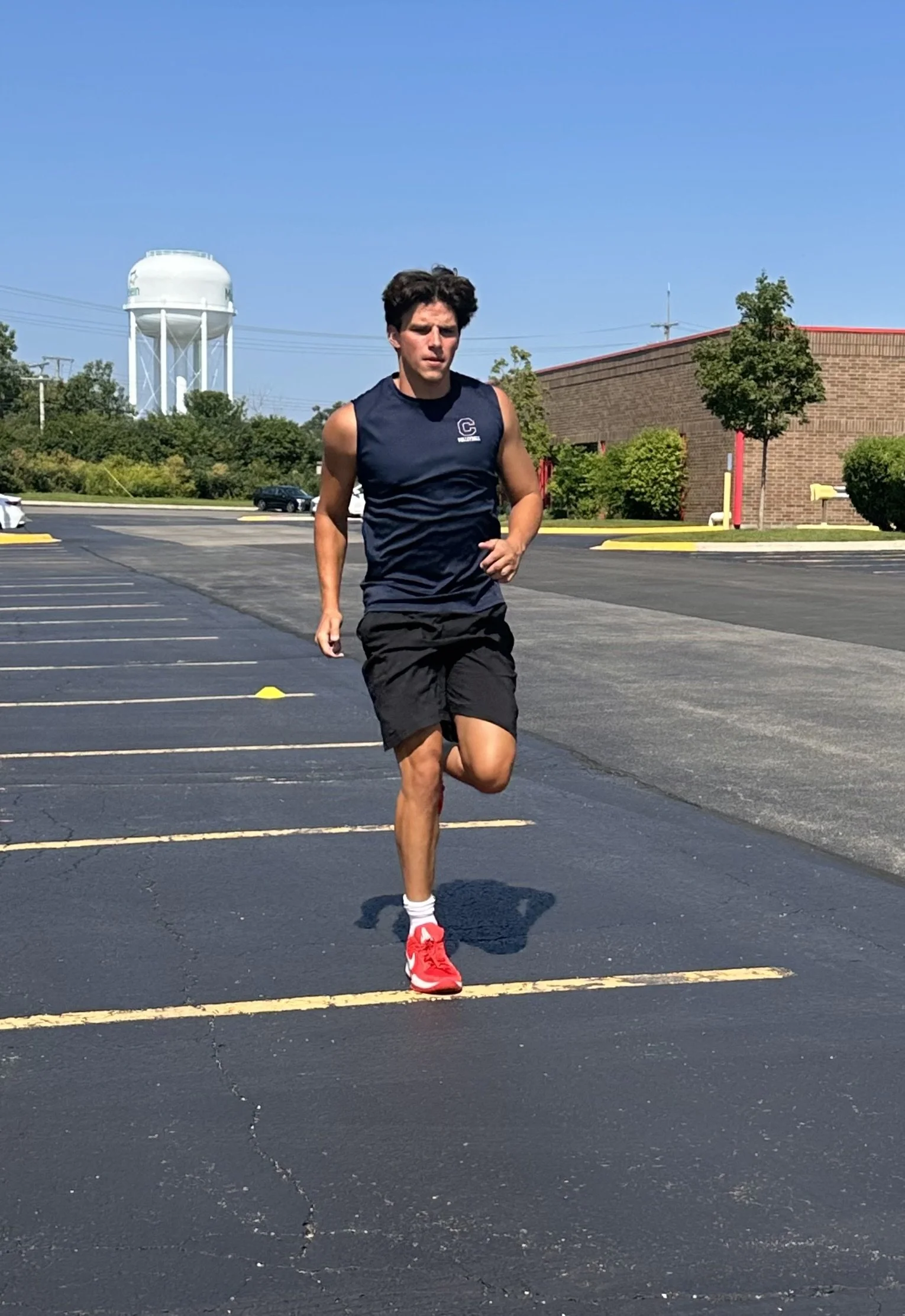 A young man with dark hair running in a parking lot on a sunny day, wearing a navy sleeveless shirt, black shorts, and red running shoes.