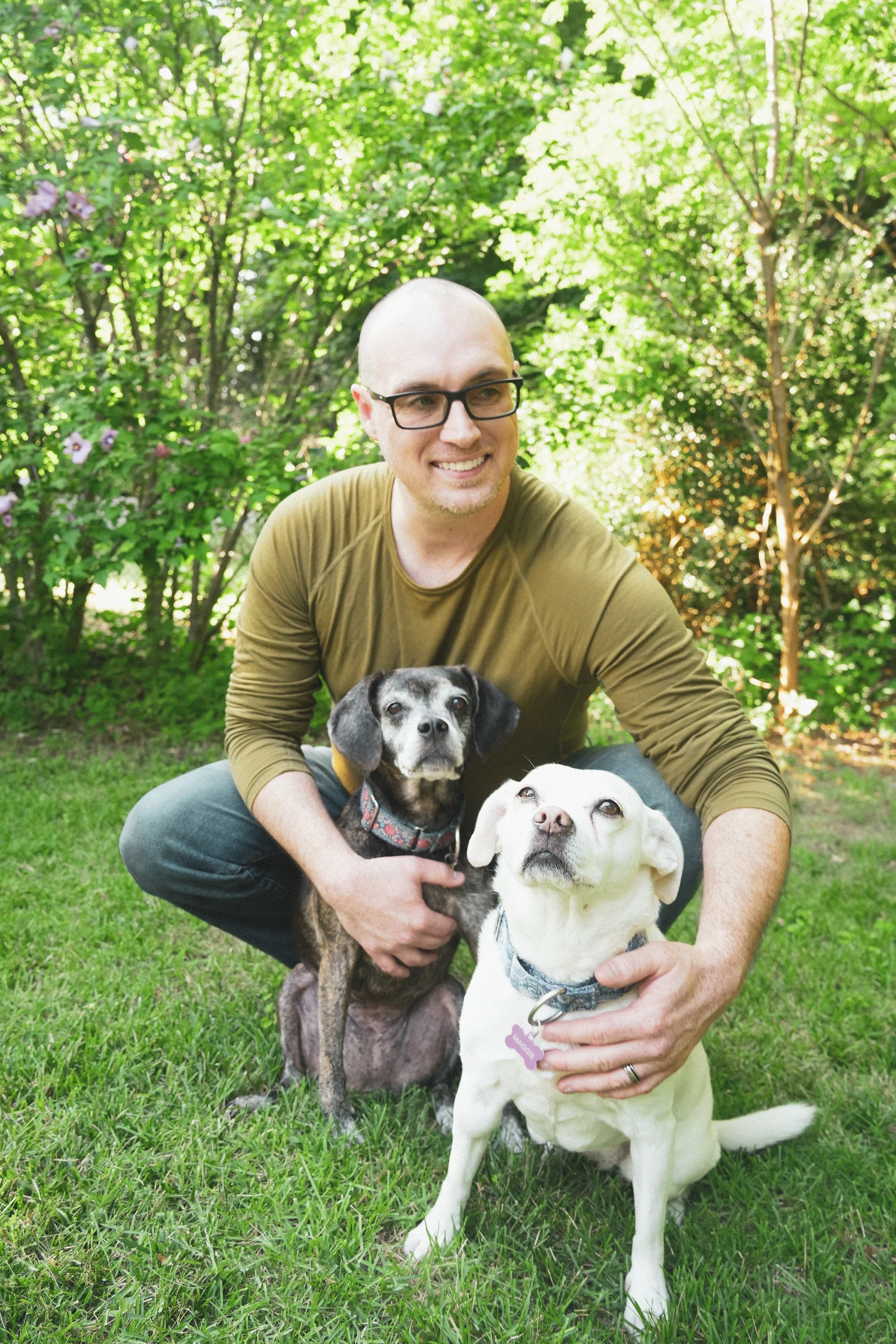 A man with glasses crouching on green grass, holding two dogs, in a background of lush trees with sunlight filtering through the leaves.