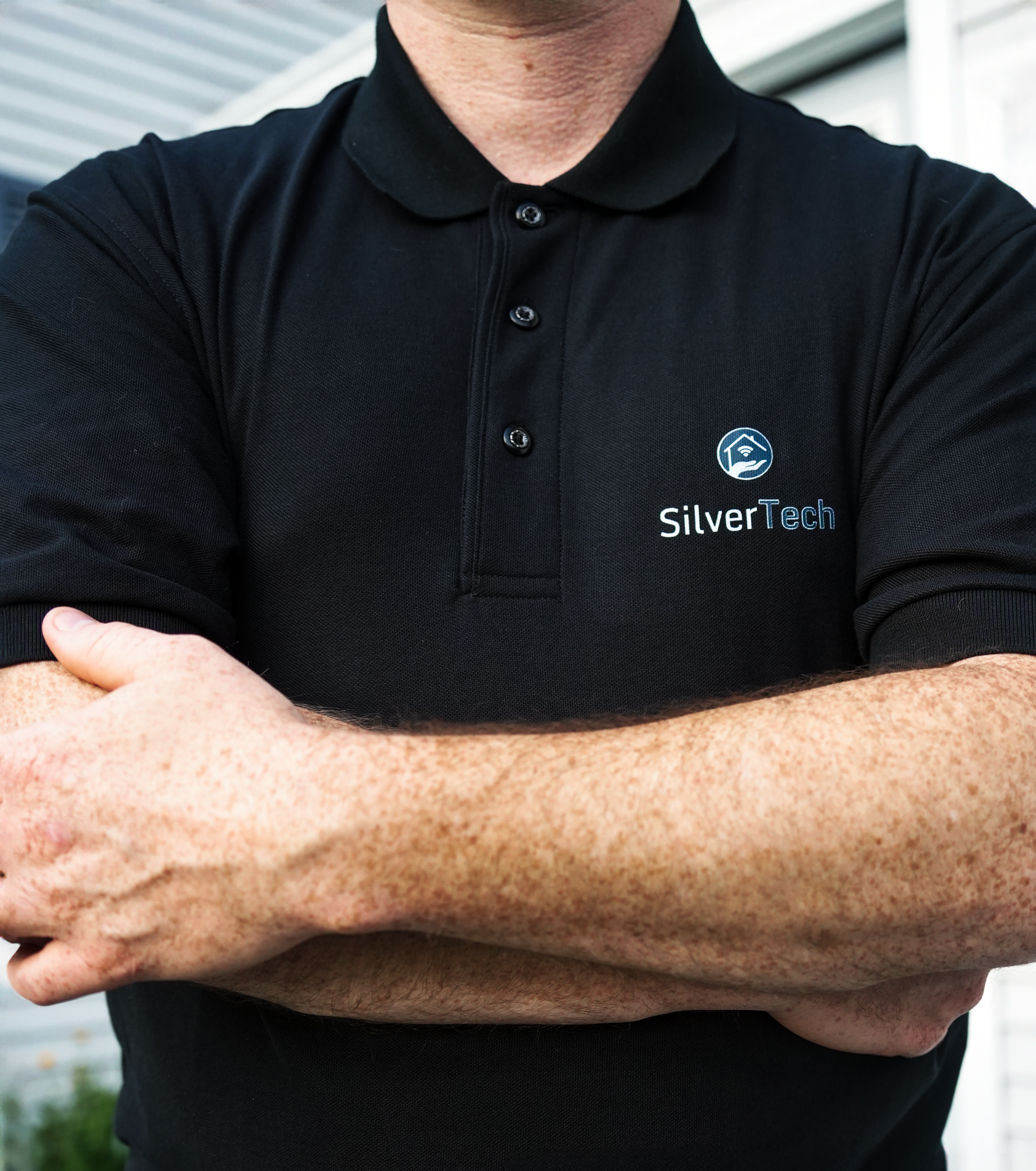 Close-up of a person wearing a black polo shirt with a logo and text that reads SilverTech, standing outdoors near a building with siding. The person's face and upper body are visible, with arms crossed and one hand resting on the forearm.