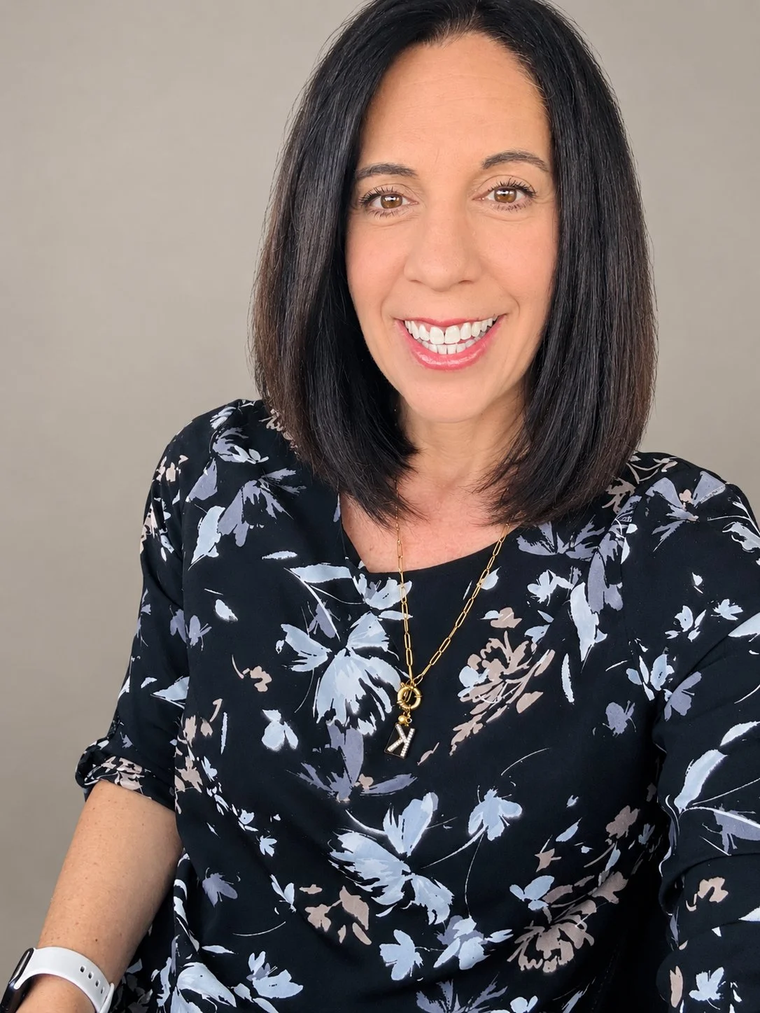 A woman with shoulder-length dark hair, smiling, wearing a black floral top and gold necklace with a "Y" pendant, against a light gray background.