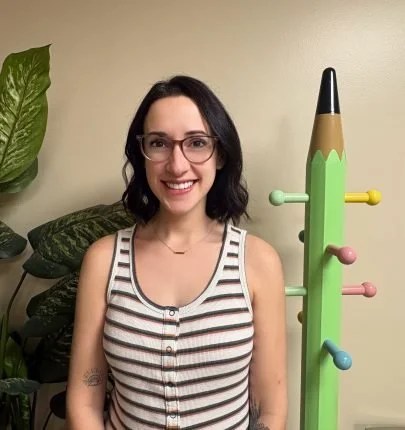 A woman with shoulder-length dark hair and glasses smiling, wearing a striped tank top, standing next to a colorful coat rack shaped like a cactus with pink, blue, yellow, and green knobs, in a room with a plant in the background.