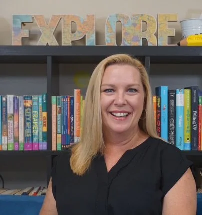 Smiling woman with blonde hair standing in front of a bookshelf with colorful books and large decorative letters spelling 'EXPLORE'.