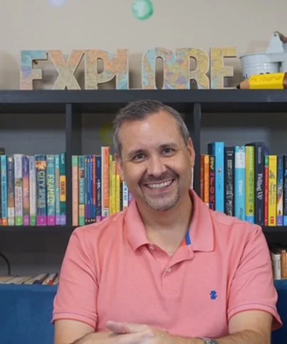 A smiling man in a pink polo shirt sitting in front of a bookshelf filled with colorful children's books. There are decorative letters spelling 'EXPLORE' on a shelf behind him.