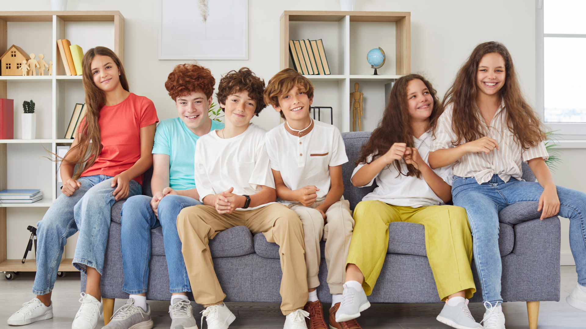 Group of six smiling children sitting on a gray couch in a bright, modern living room, with books, a globe, and decorative items on white shelving behind them.