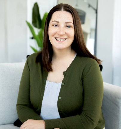 A woman with long brown hair wearing a green cardigan and light blue top, smiling, sitting in a bright room with a plant in the background.