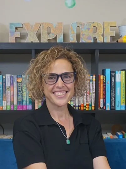 A woman with curly blonde hair and glasses smiling in front of a shelf filled with colorful books and an "EXPLORE" sign in decorative letters.