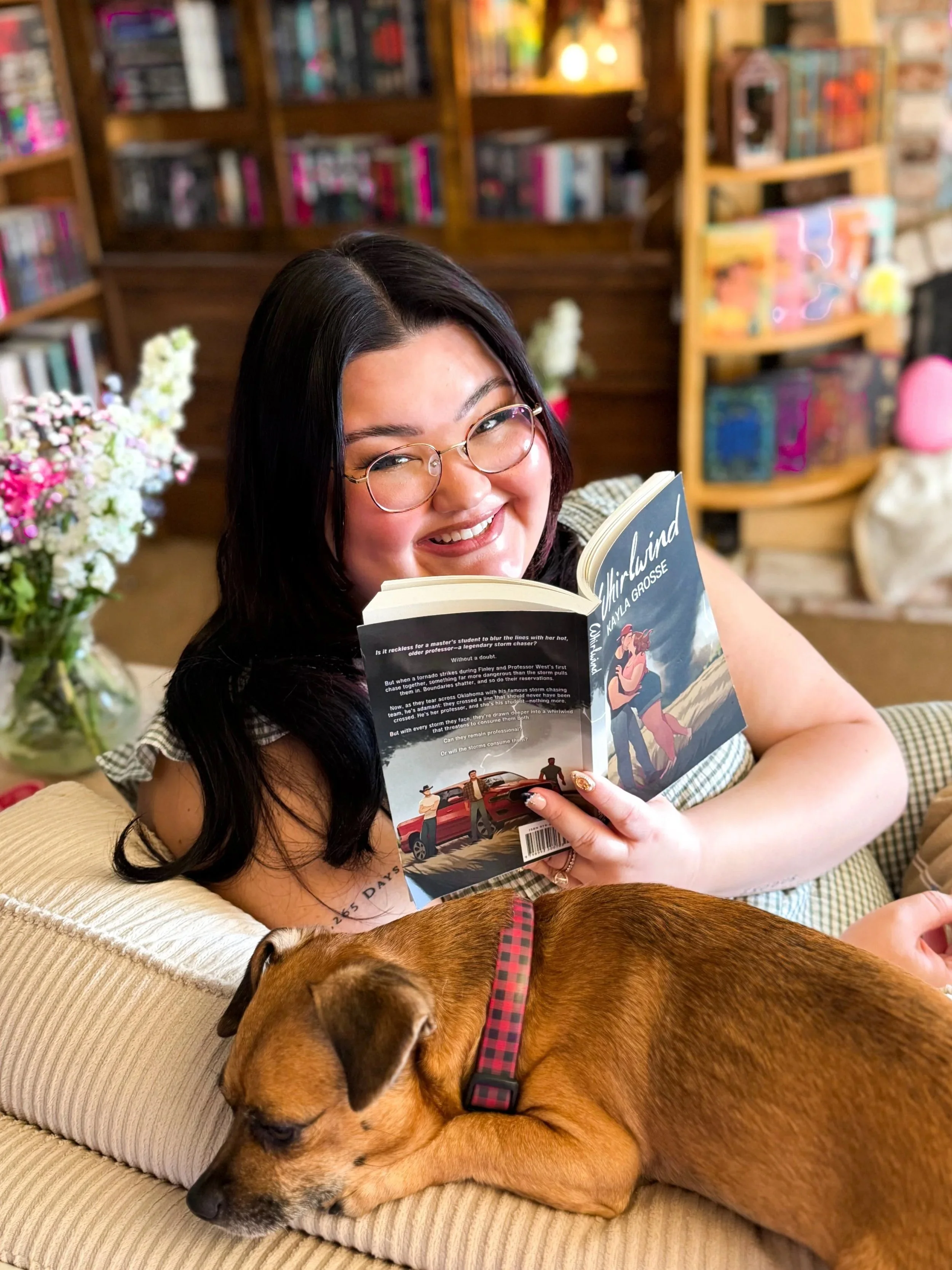 A woman with glasses and dark hair smiling while reading a book, a brown dog with a red and black collar sleeping on a beige couch in front of her, a bookshelf with books and decorations in the background, and a vase of flowers on the side.