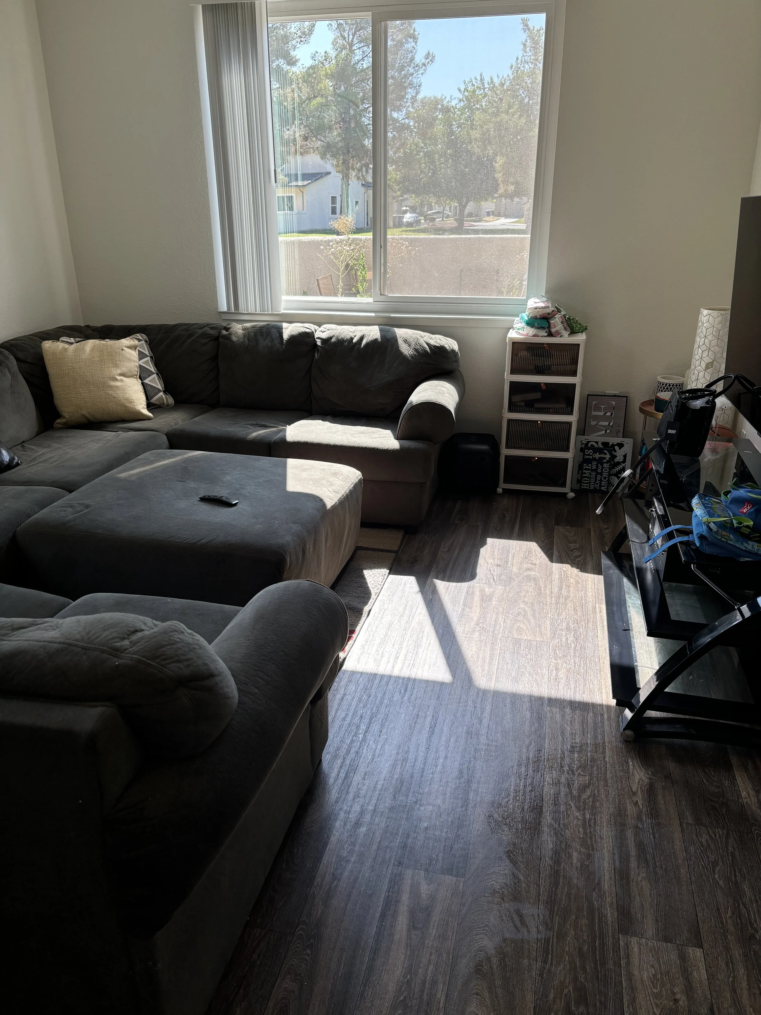 Living room with a large window, a gray sectional sofa with pillows, a small white storage drawer, and a TV stand with assorted items on it. The sunlight casts shadows on the dark wood floor.