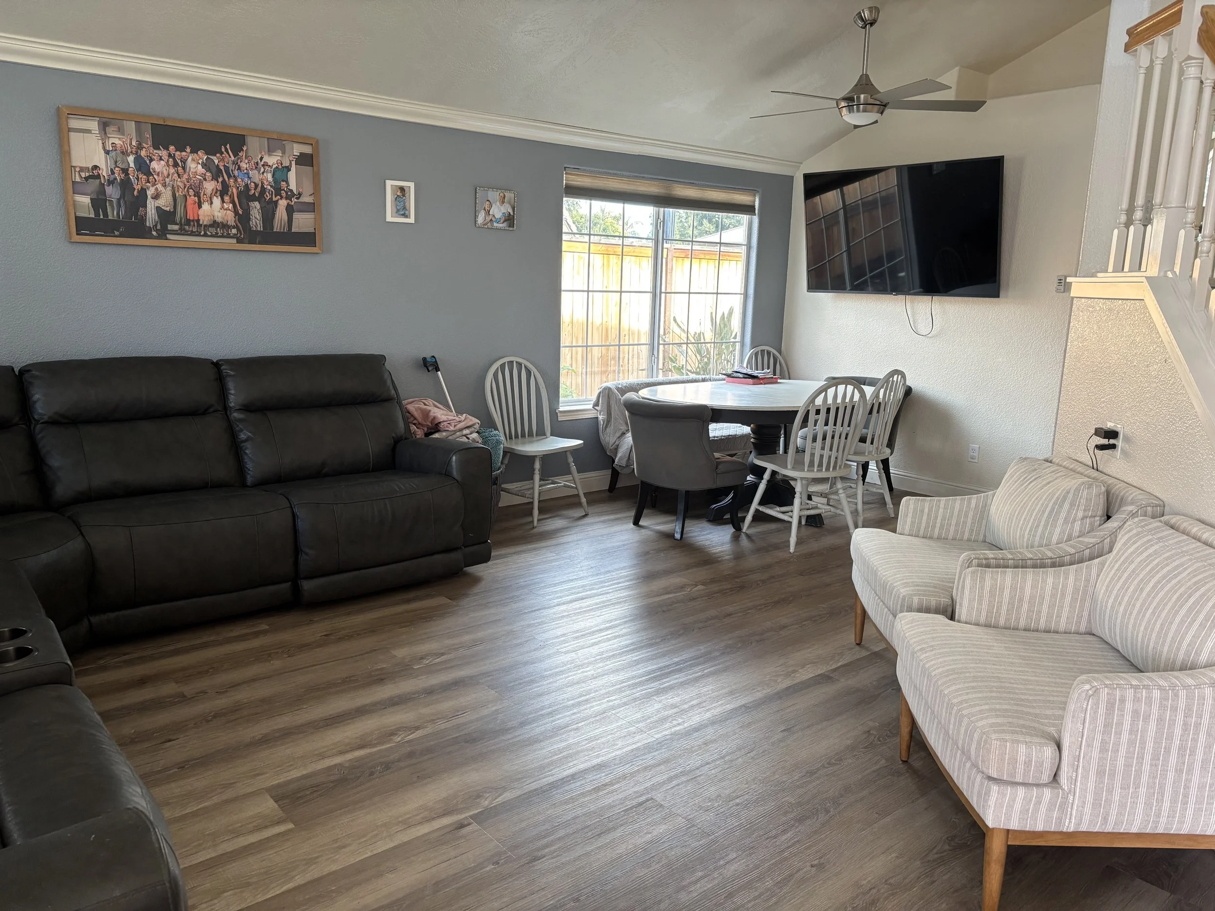 A living room with a black sofa, a striped armchair, a dining table with four white chairs, a large flat-screen TV on the wall, and three framed photos on a blue wall, with a window and ceiling fan.