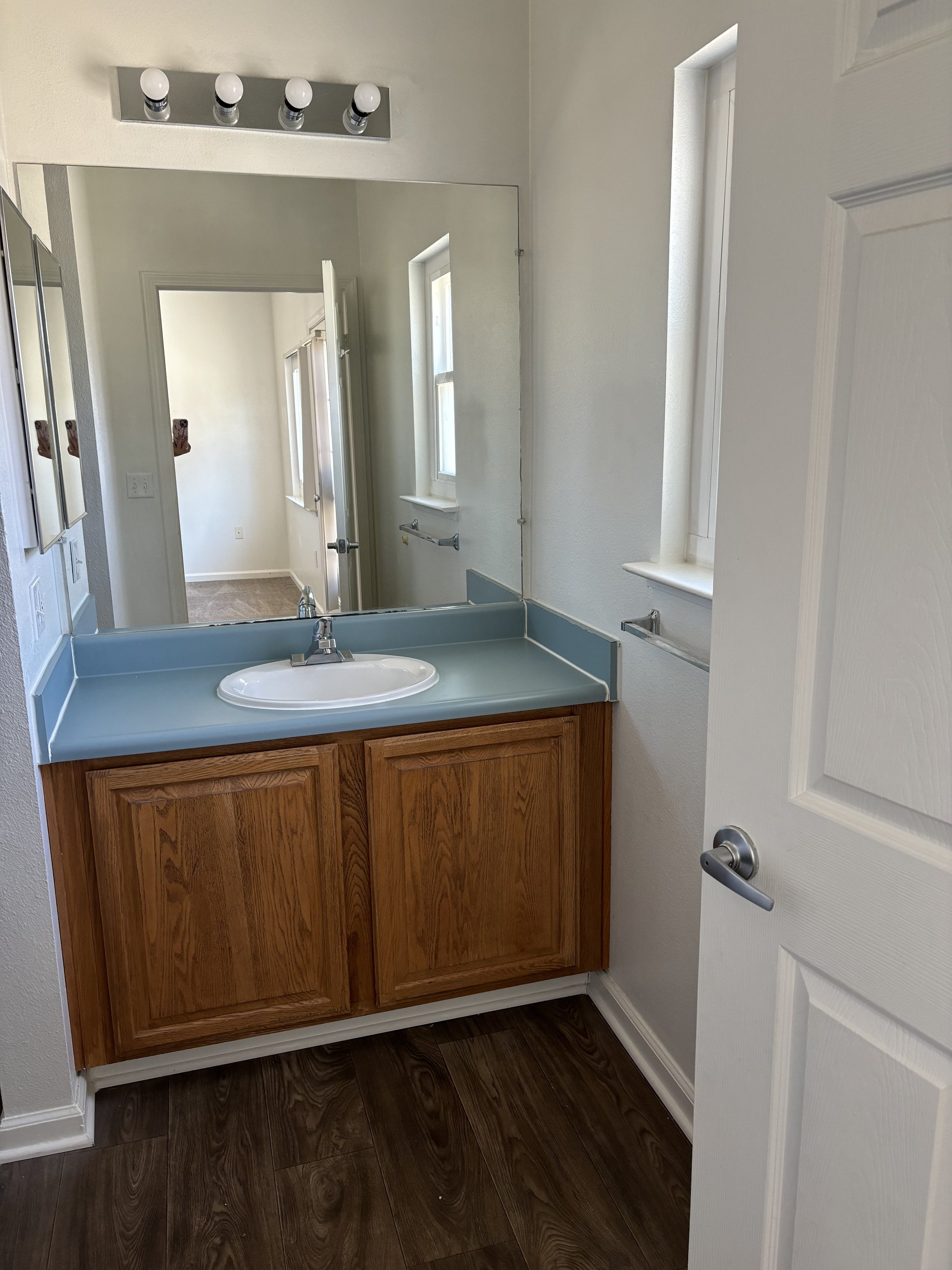 A bathroom with a wooden vanity, a mirror, a sink, and a light fixture with four bulbs. The bathroom has white walls, two narrow windows, and a door. The floor is dark wood.