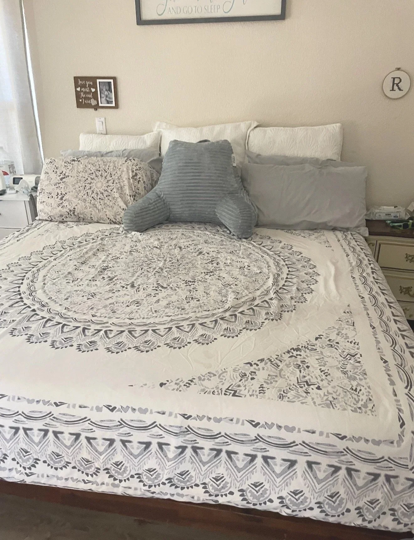 A neatly made bed with a white and gray patterned bedspread, five pillows in white and gray, and a gray plush bear in the center. There are two nightstands with various items on either side of the bed, and framed wall art hanging above.