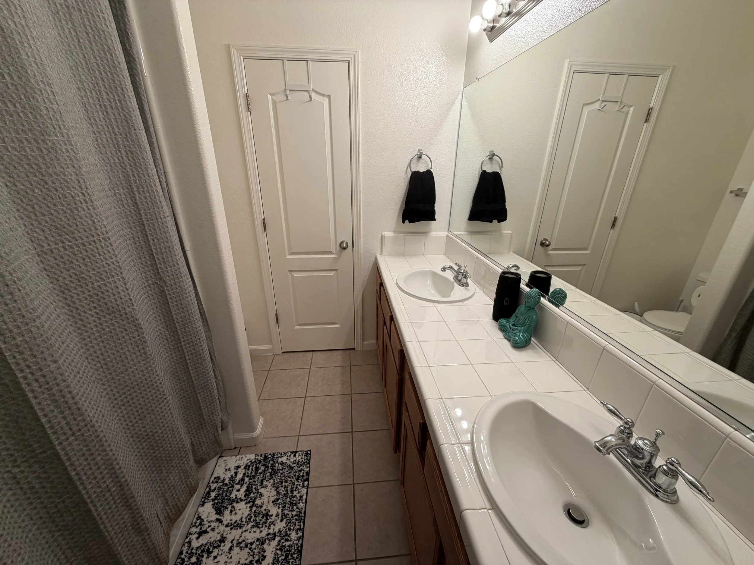 A bathroom with a white tiled countertop, a sink, a large mirror, black hand towels, and a decorative Mahjong Buddha sculpture on the counter. There is a door and a shower area with a textured shower curtain.