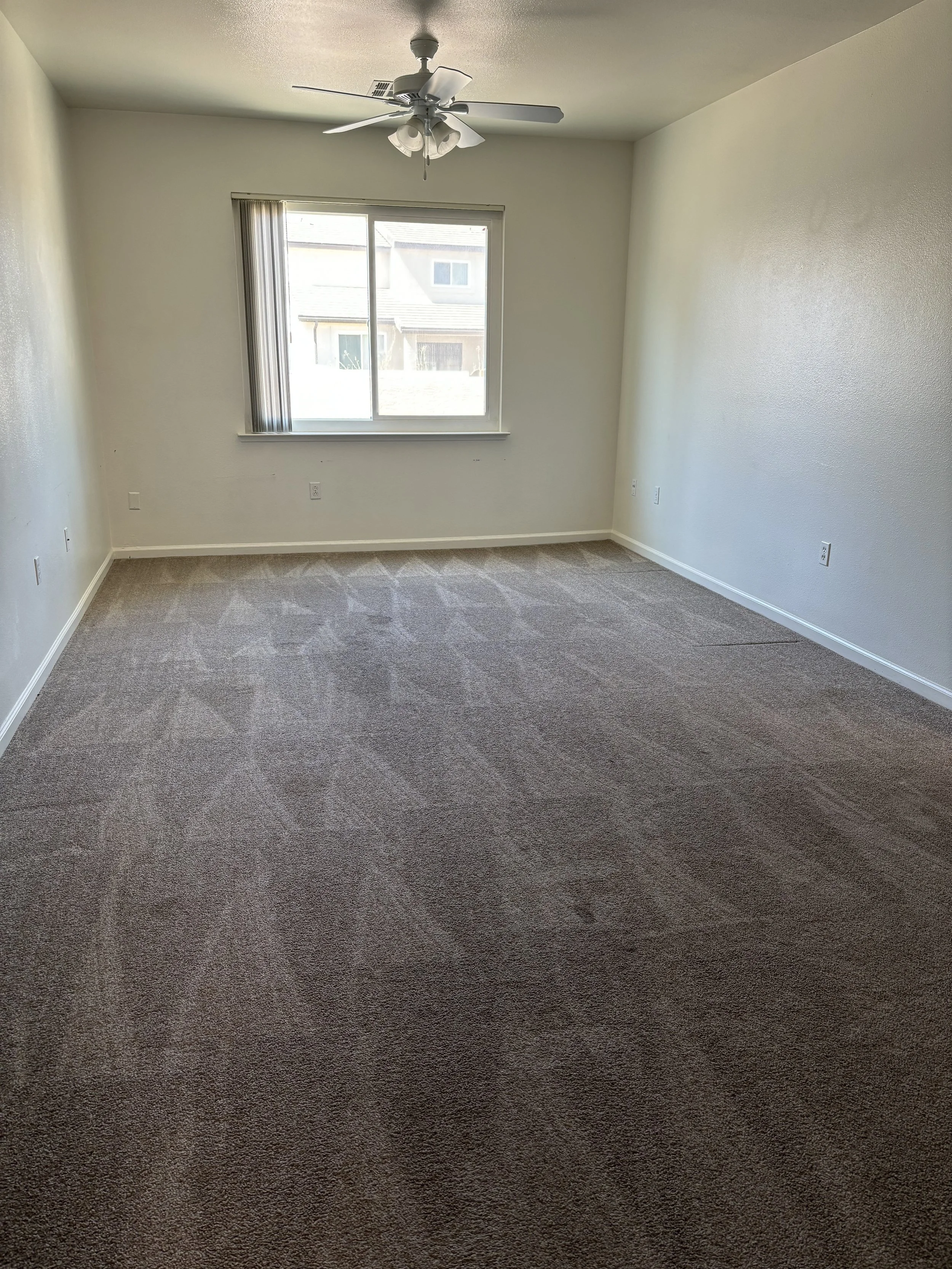 Empty room with beige carpet, white walls, a window with vertical blinds, a ceiling fan with lights, and multiple electrical outlets.