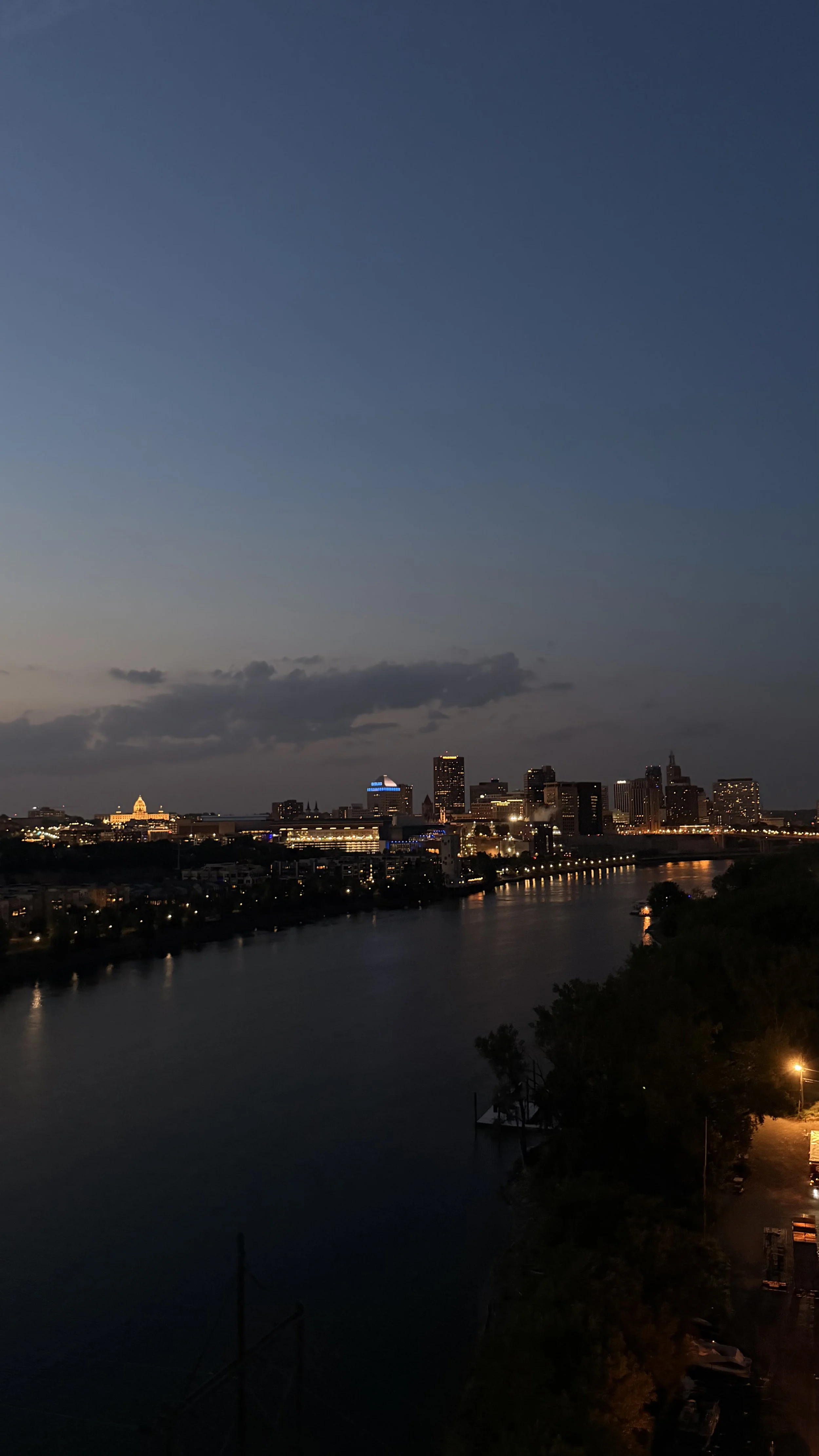 City skyline at dusk with illuminated buildings along a river, with some clouds in the sky and a tree-lined promenade on the right.