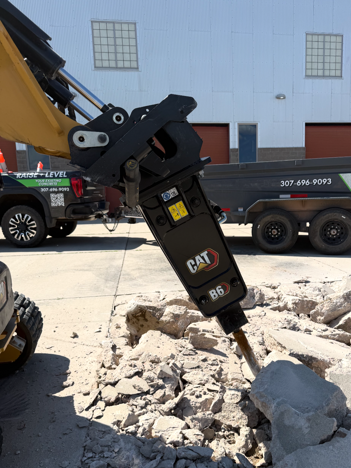 A black hydraulic attachment connected to a piece of construction equipment, placed on a wooden pallet on the ground in a construction area.