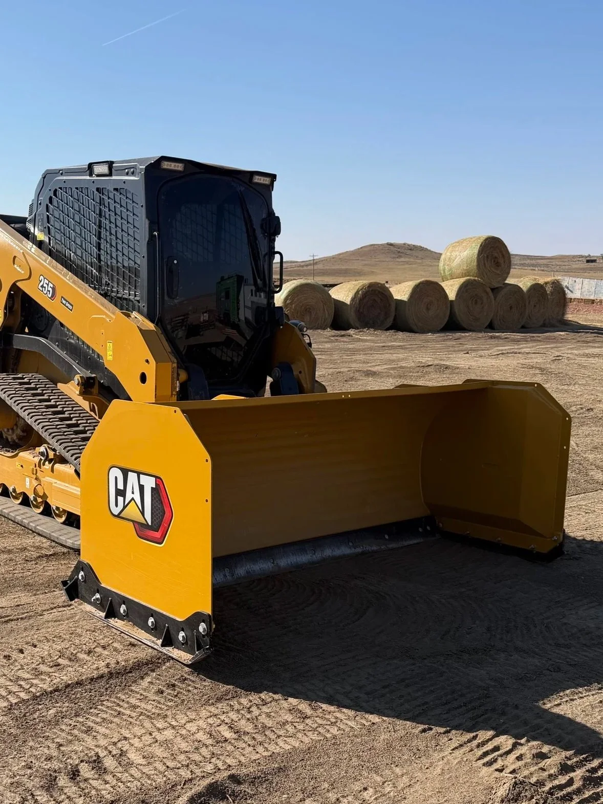 A yellow construction or agricultural machine attachment with hydraulic hoses and metal components, likely used for ground work or snow removal, situated outdoors on a dirt surface.