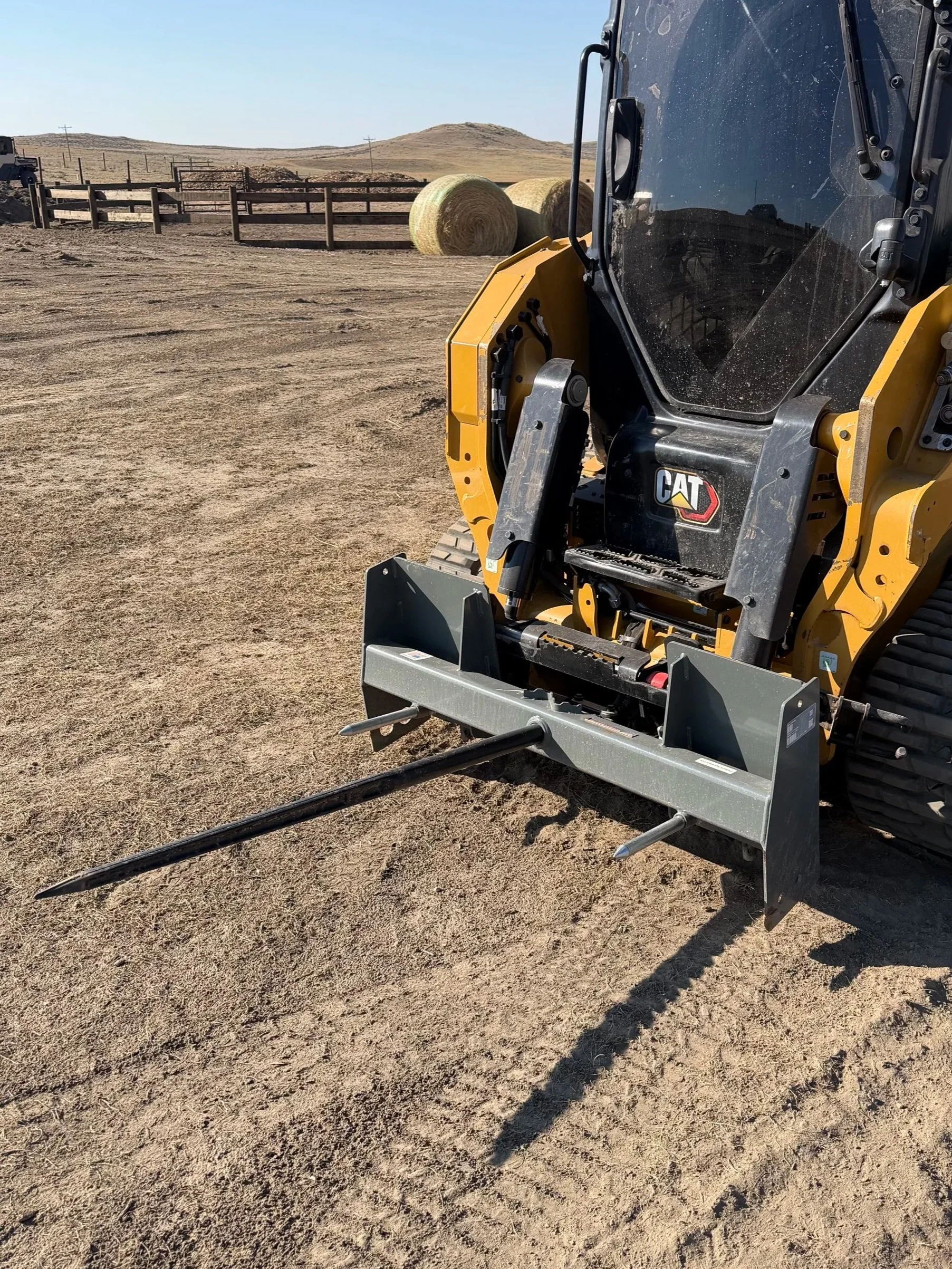 A collection of metal pipes arranged horizontally and bundled with straps. The pipes are placed on wooden supports on a wooden pallet. There is a yellow machine in the background and a gray metal device on wheels in the foreground on a dirt surface.