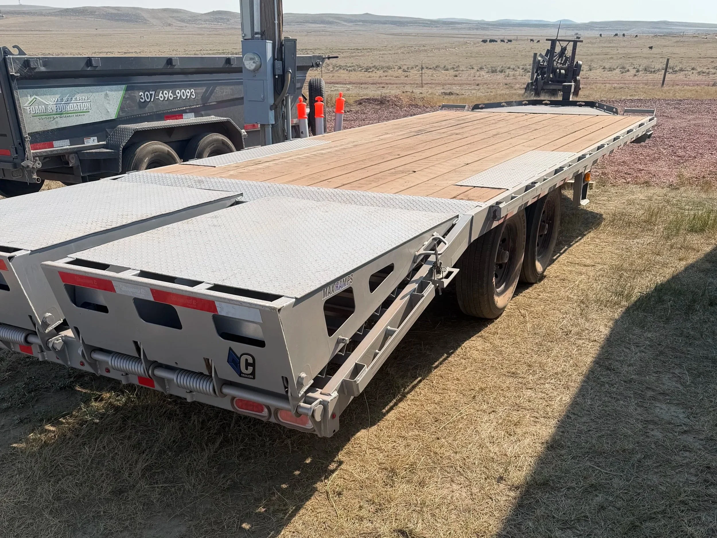 A flatbed trailer with a wooden floor is parked on a grassy field, with a large machinery or equipment in the background and wide open plains in the distance.