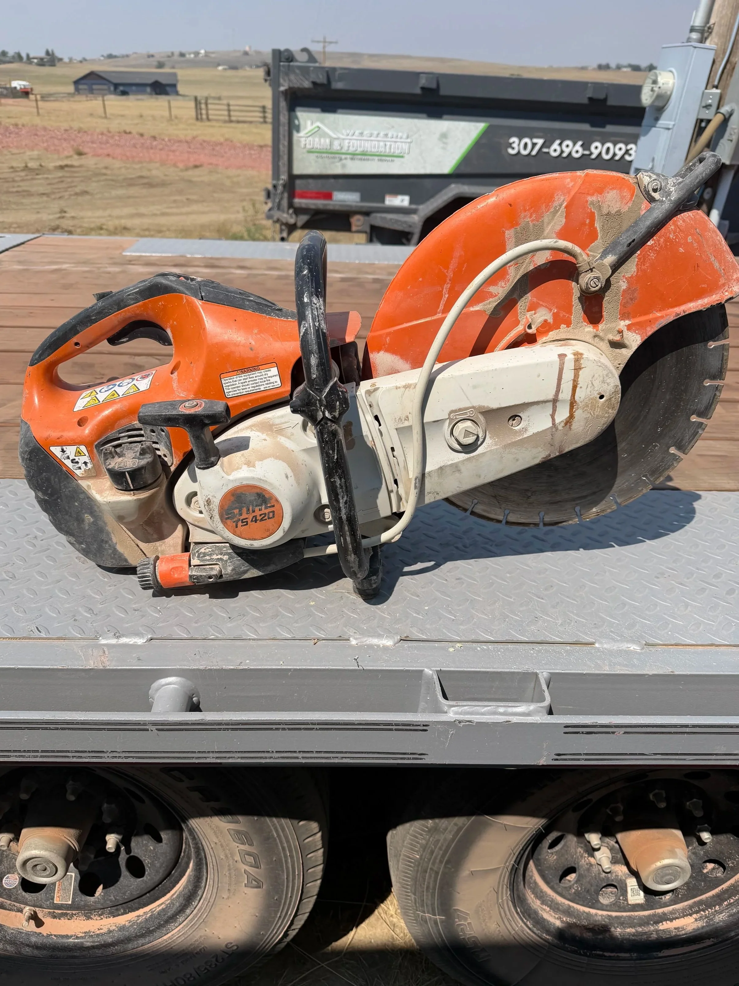 Close-up of a portable concrete saw with orange and white body, mounted on a metal surface, with a large saw blade and a power cord, with a trailer and a rural landscape in the background.