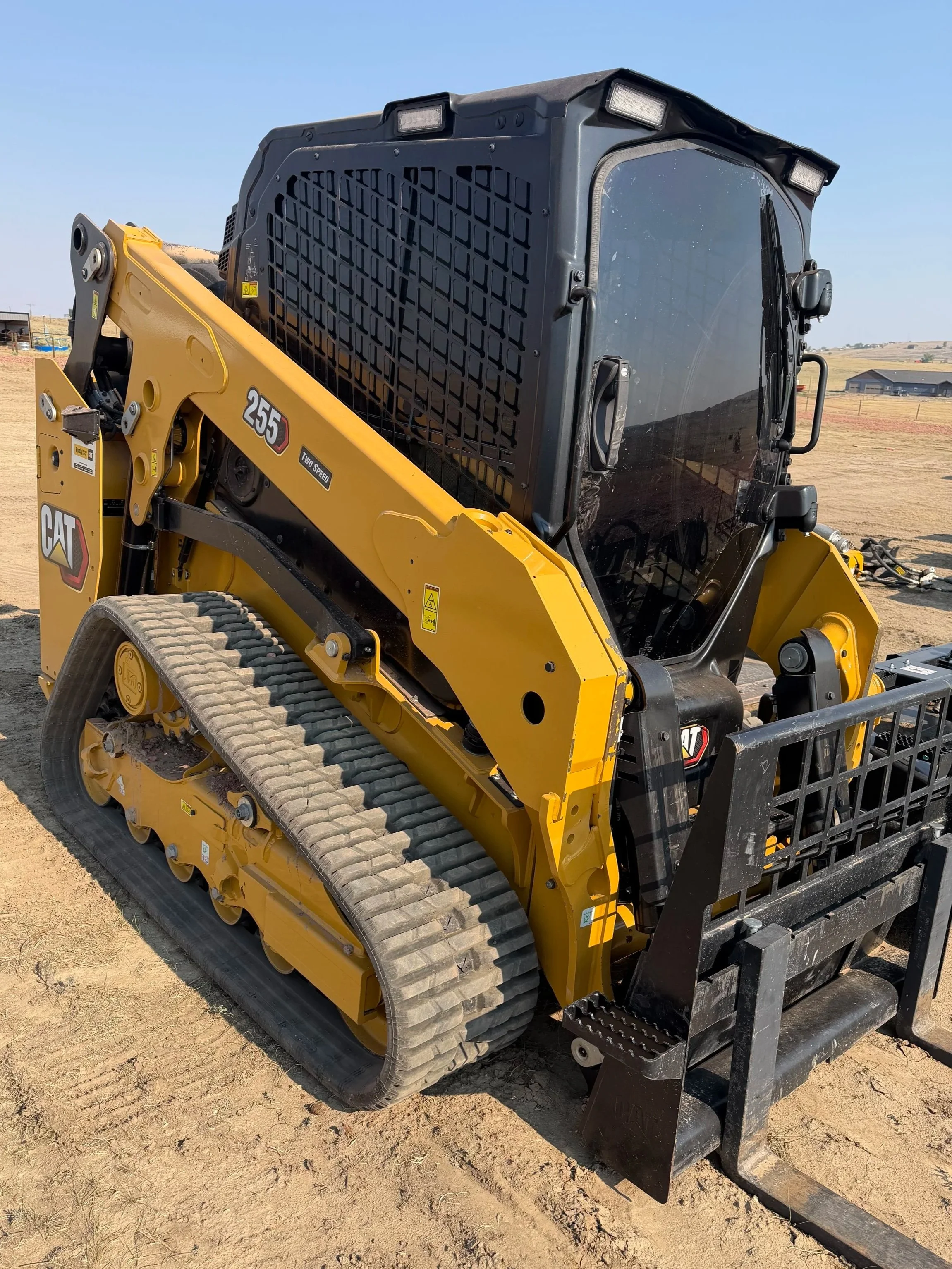 A yellow and black Caterpillar compact track loader with an enclosed cab, sitting on dirt in an open area with clear blue sky and distant buildings.