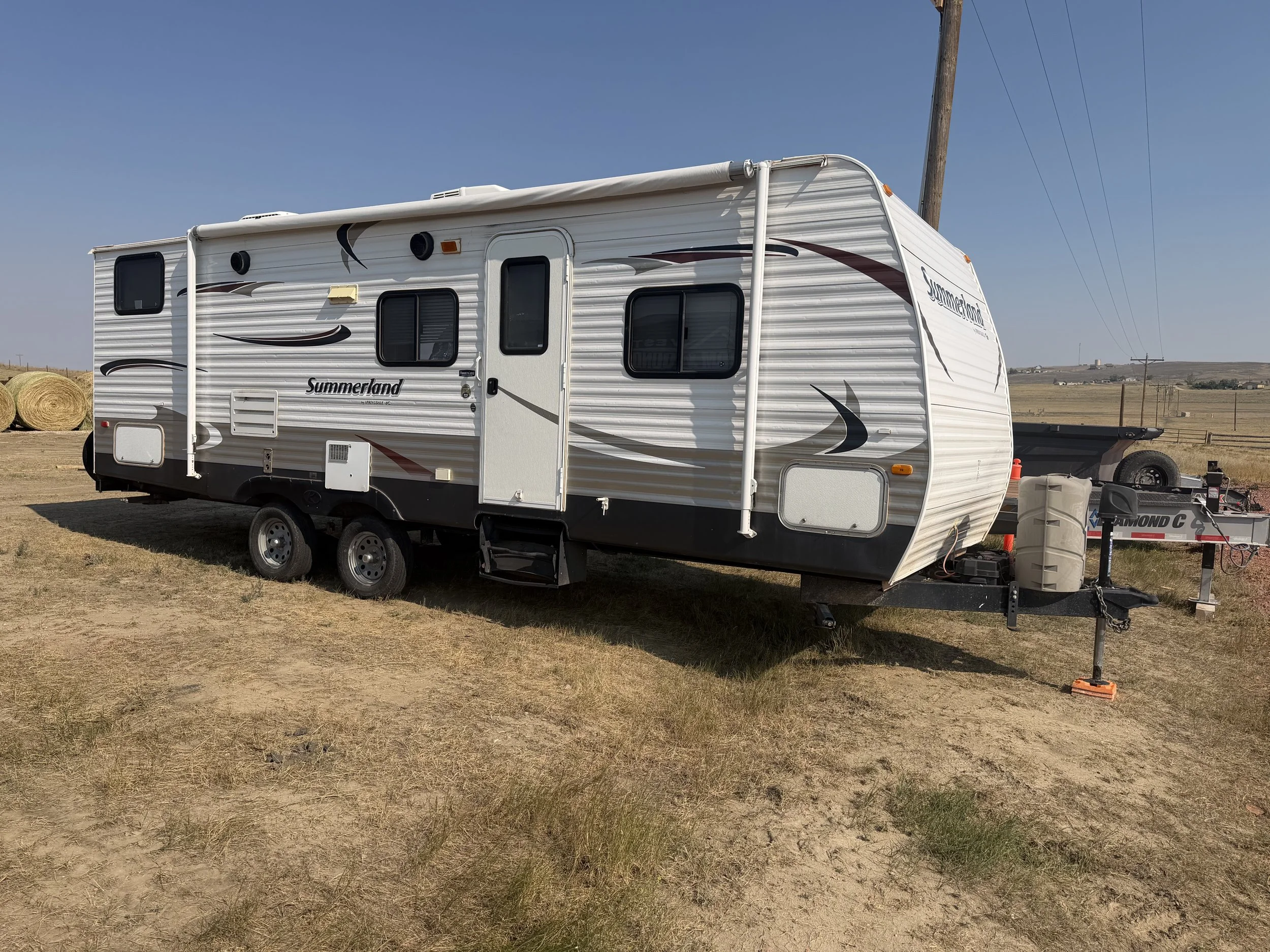 A white travel trailer with black and maroon graphics parked on dirt with hay bales nearby and a utility trailer attached, under a clear blue sky.