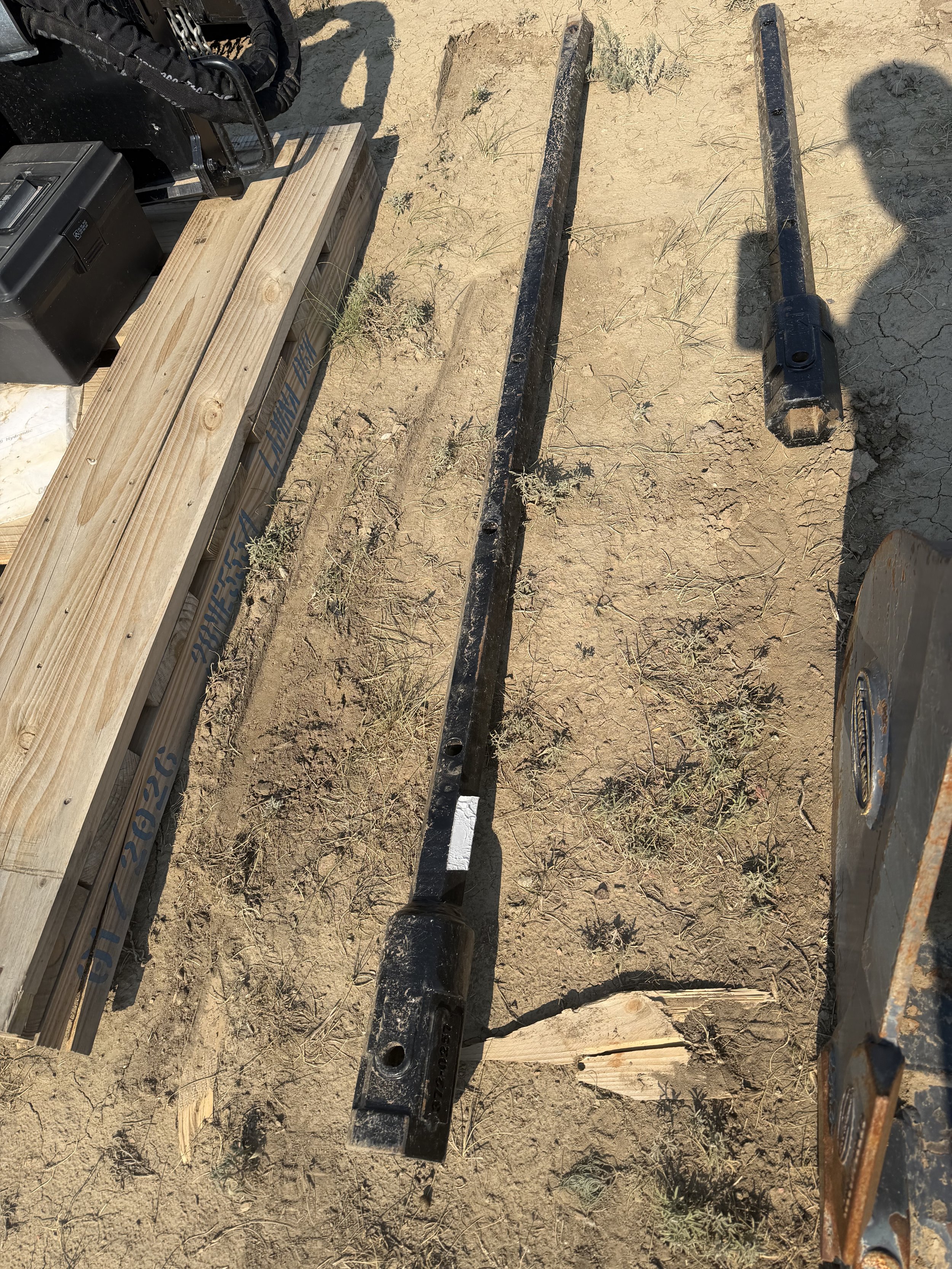 Two long metal bars or rods laid parallel on dry, cracked dirt with sparse small plants. Part of a wooden structure and some equipment are visible on the left side of the image.