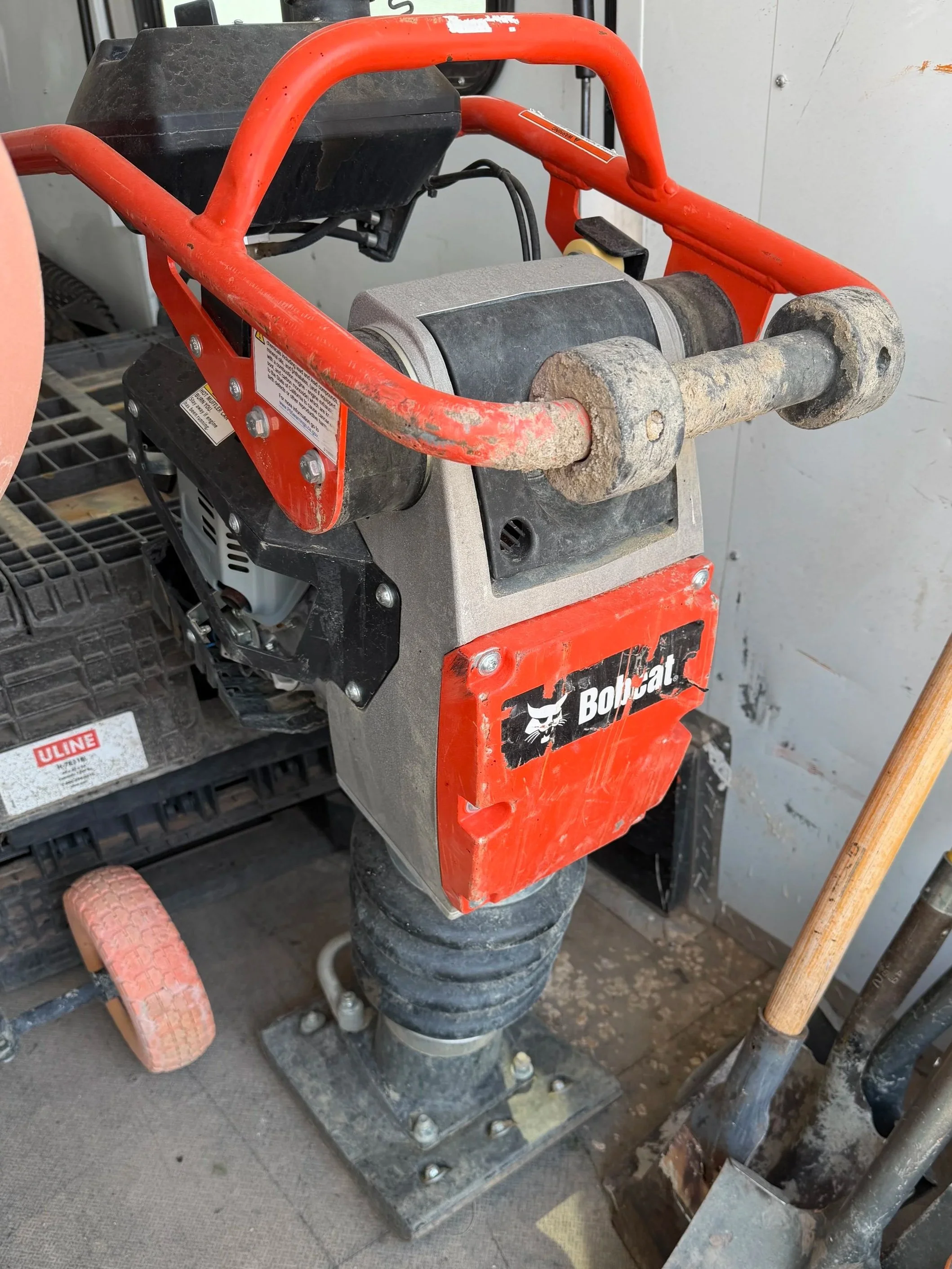 Construction equipment with a red frame, black motor, and a large cylindrical roller attached to the bottom, surrounded by a shovel and tools in a workshop.