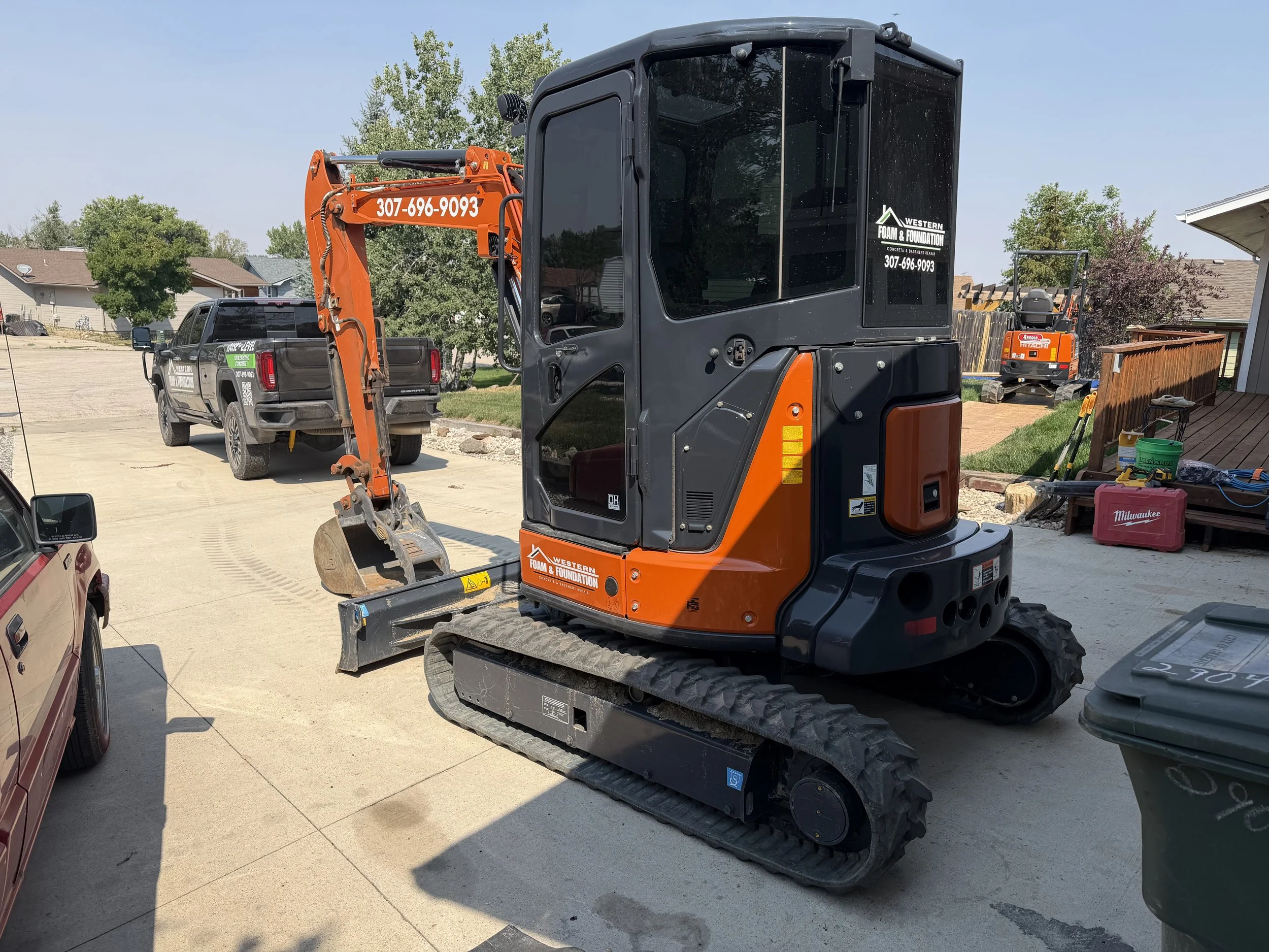 Small orange and black excavator with roofing company branding, parked in a residential driveway, with a pickup truck and other construction equipment visible in the background.