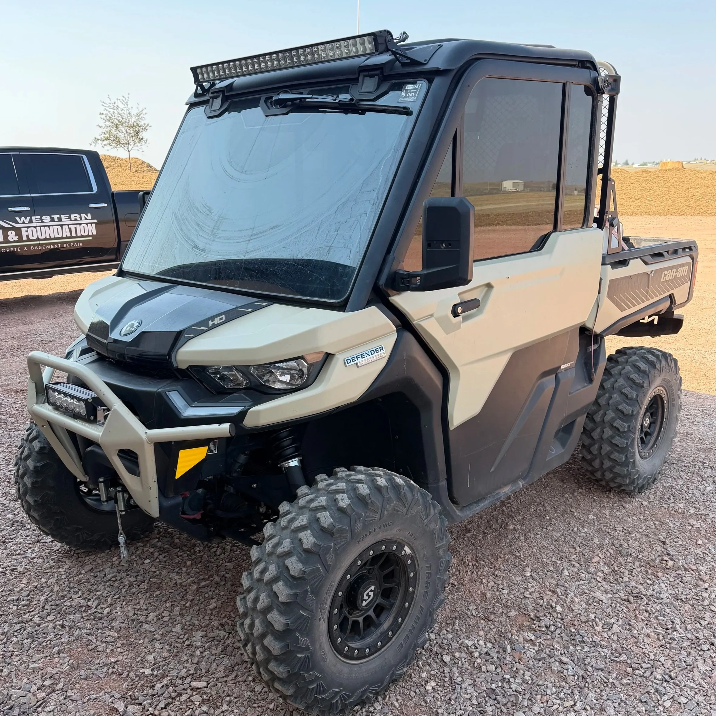 A close-up of a white off-road utility vehicle with large, rugged tires and a black roof, parked on gravel with a construction site and a tree in the background.