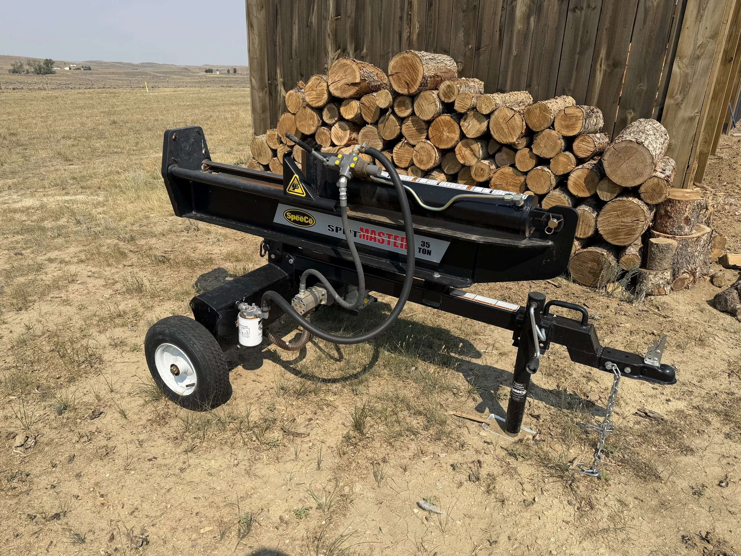 A wood splitter with a 35-ton capacity, positioned outdoors on a grassy field near a pile of cut logs, with a wooden fence and open landscape in the background.