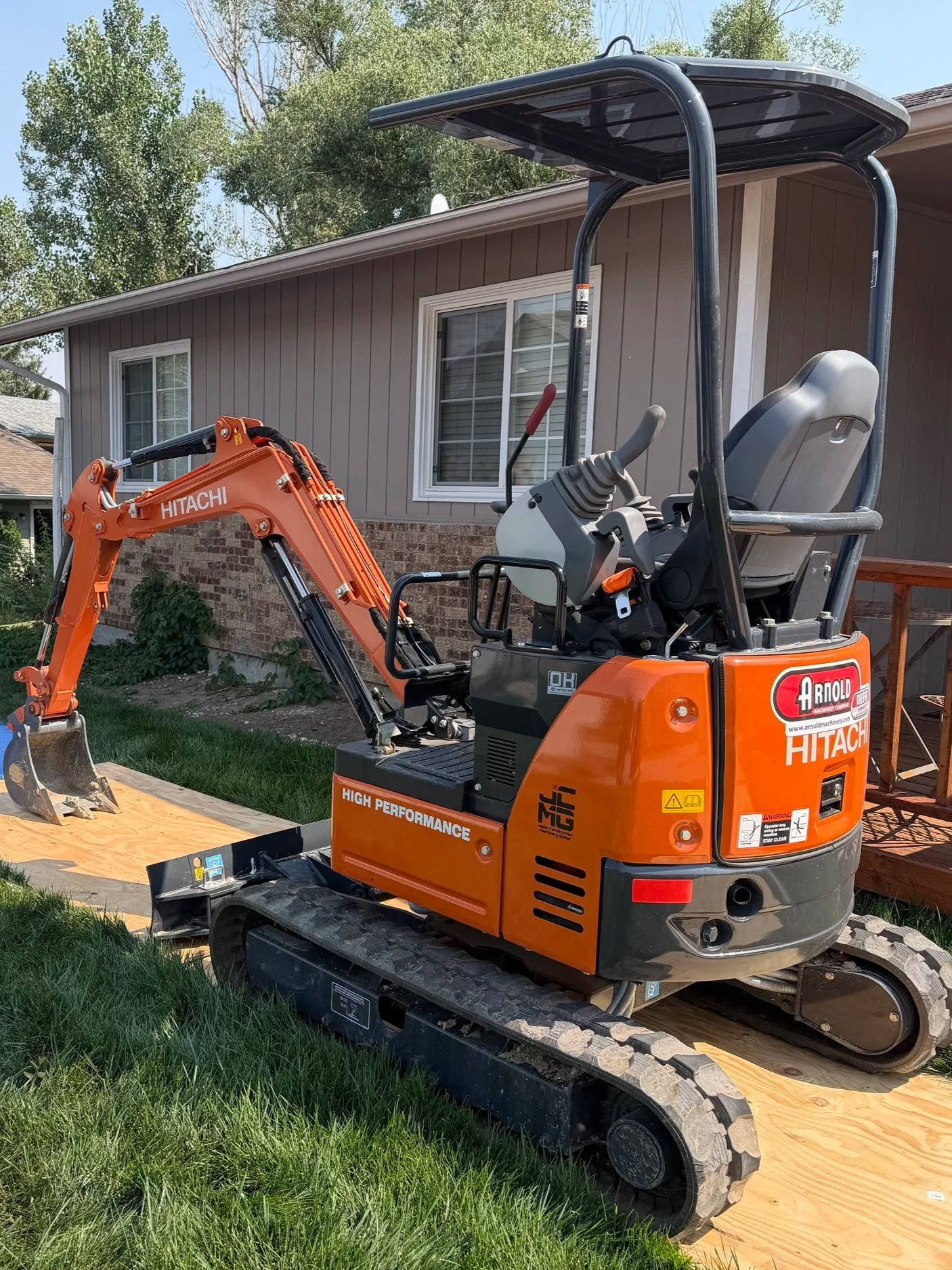 A small orange and black mini excavator with a back canopy parked on a wooden board in a backyard near a gray house with white window frames and green trees in the background.
