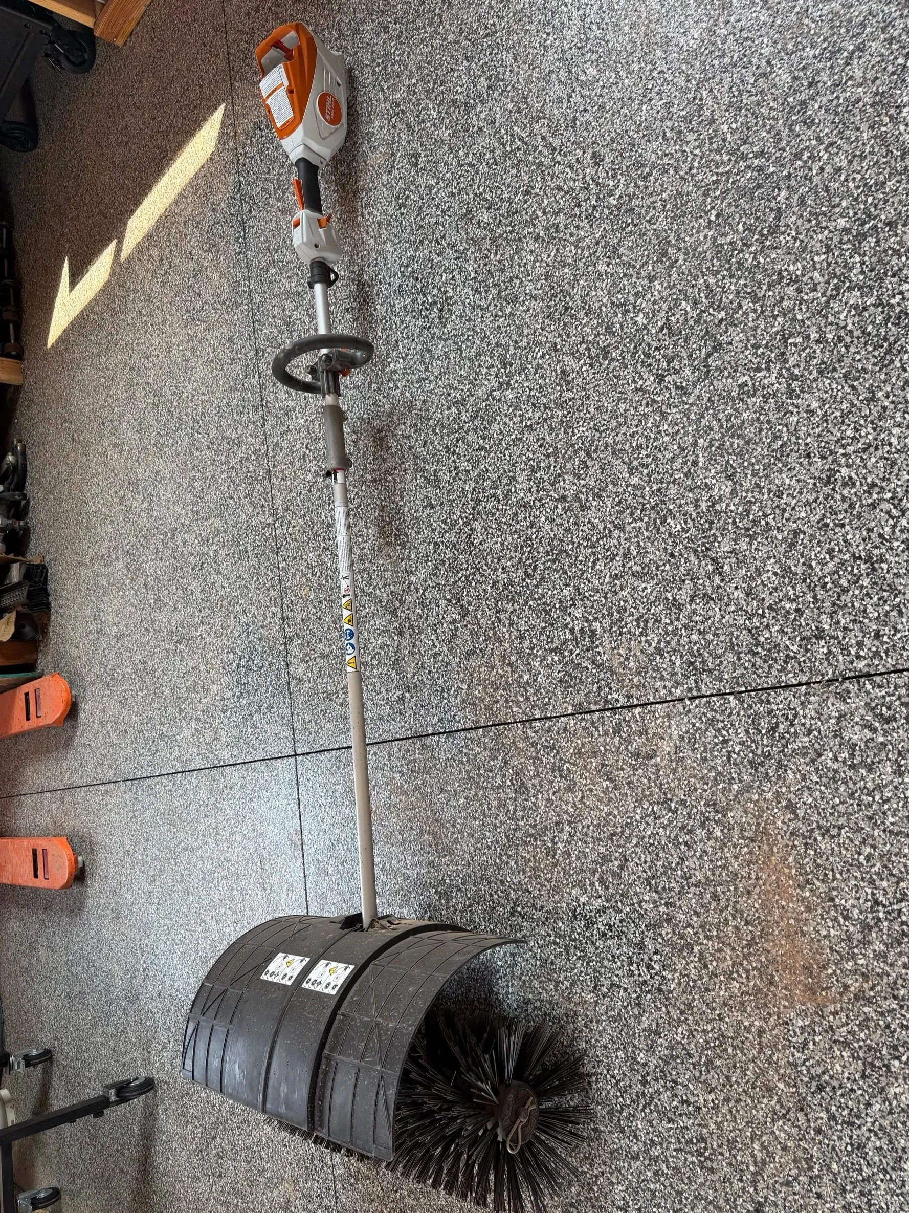 A street sweeper attachment with a cylindrical spinning brush and a long handle, lying on a speckled gray floor in a garage or workshop setting.
