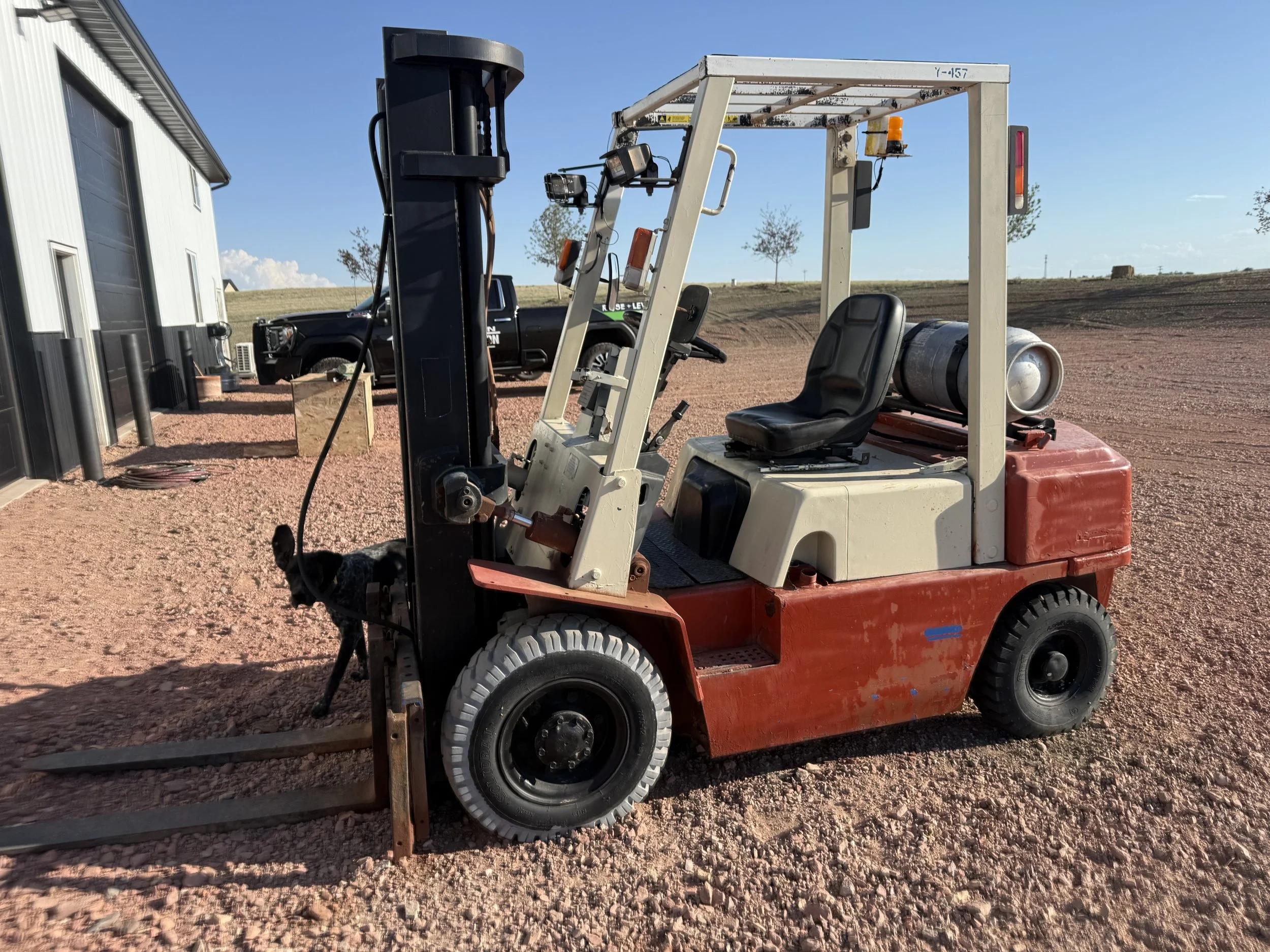 A small red and white forklift with a black seat, parked on a gravel surface outdoors with a clear blue sky, a black truck, and a white building in the background.