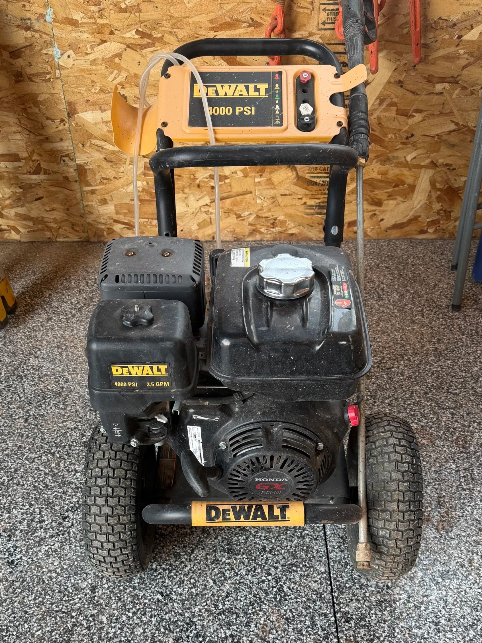 A portable DeWalt air compressor with a black Honda gasoline engine, placed on a speckled garage floor with plywood wall in the background.