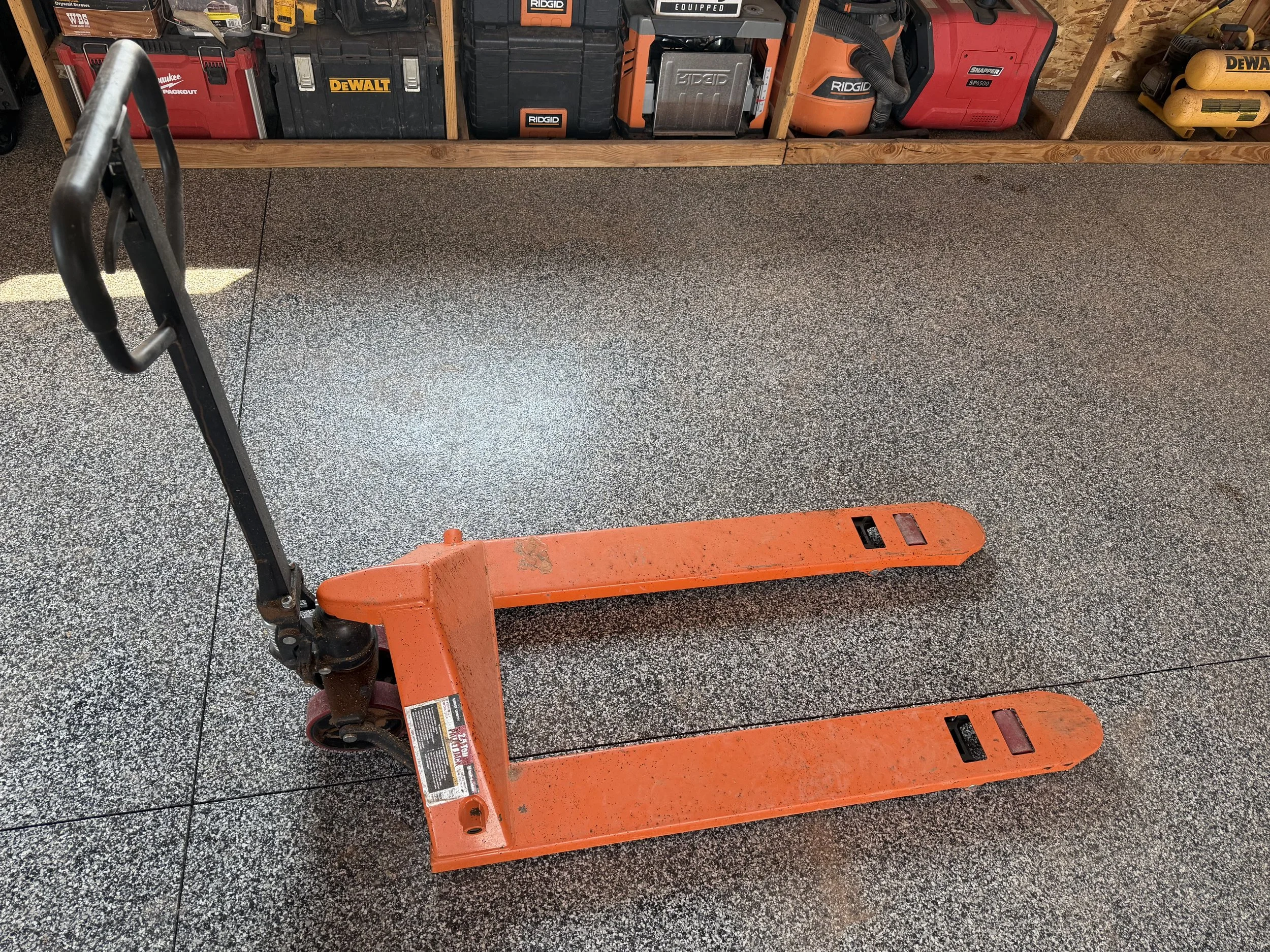 A garage workspace with textured gray flooring and wooden shelving holding power tool cases. An orange pallet jack is in the foreground, partially visible.