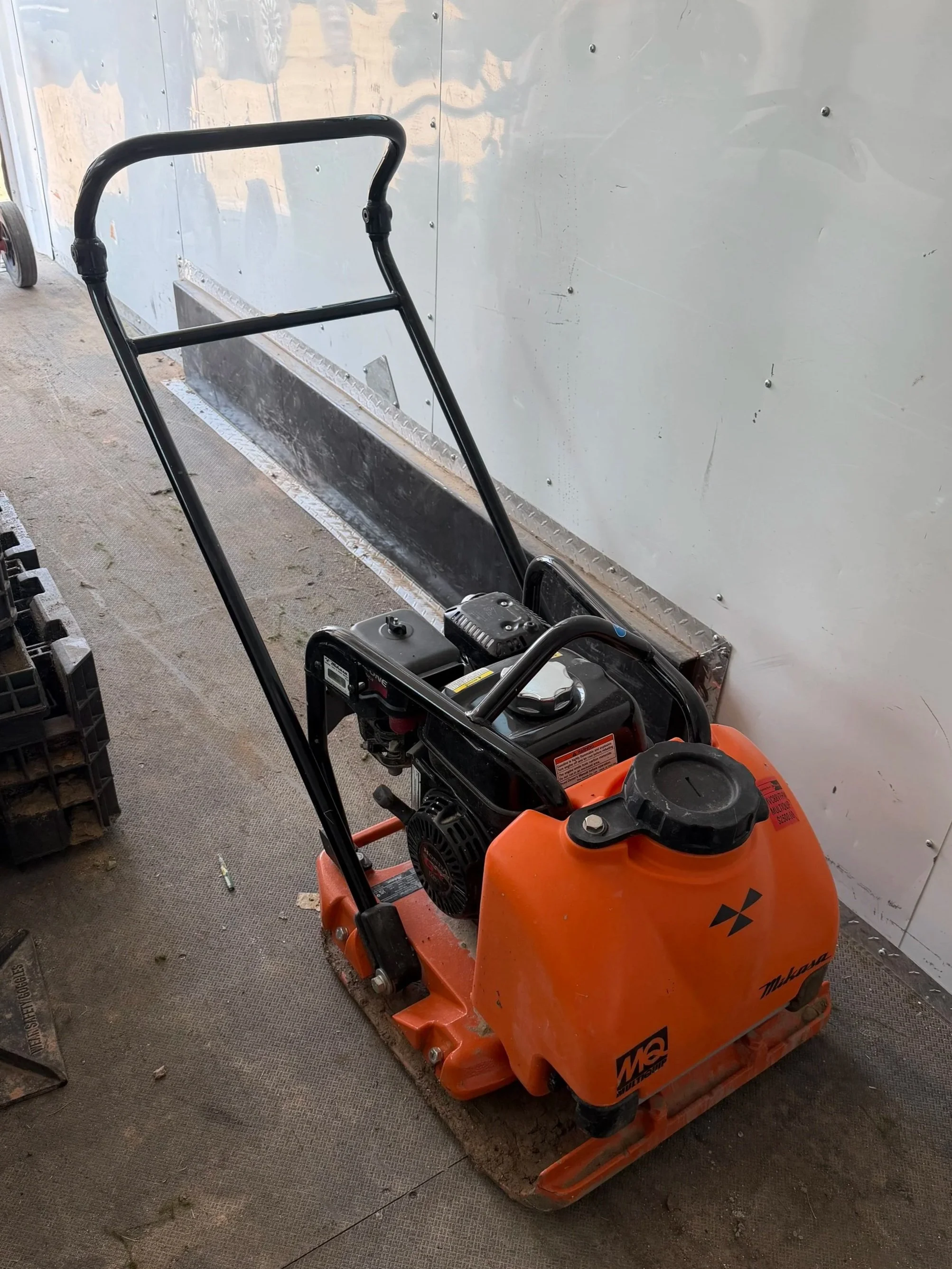 An orange commercial floor grinder with a black handlebar and engine, placed on a wooden pallet on a concrete floor, with a white wall in the background.