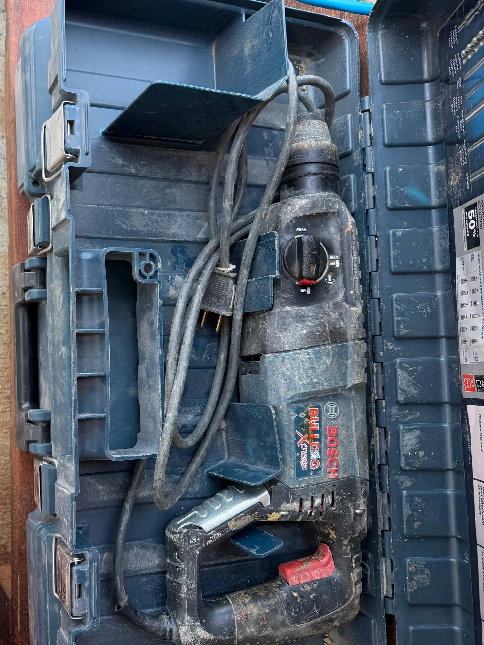 A Bosch cordless drill with a black and red handle and a silver drill bit, sitting inside an open blue plastic toolbox with a Bosch label on the side.