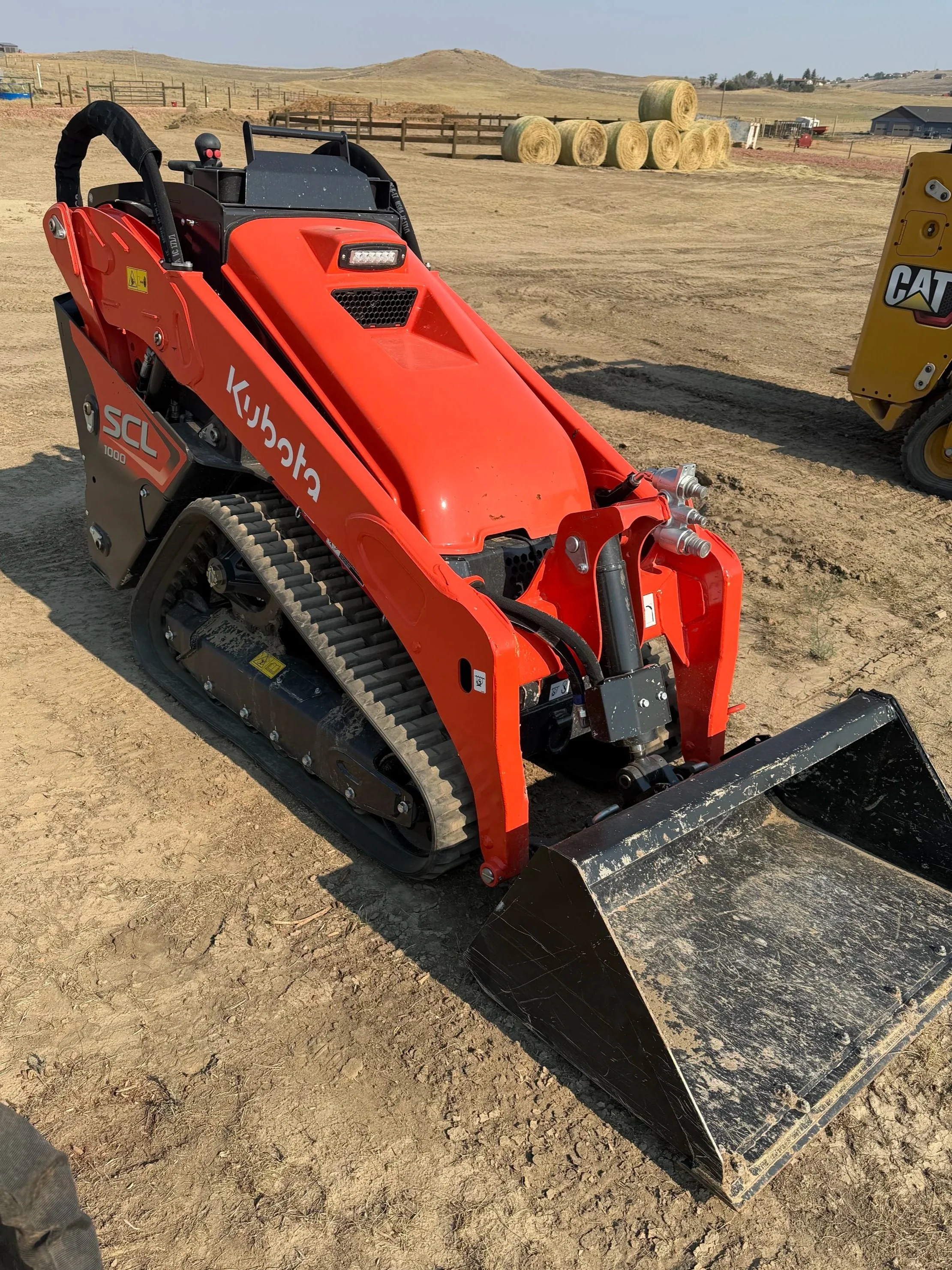 A red compact tracked loader on a dirt field with hay bales in the background and a rural landscape.